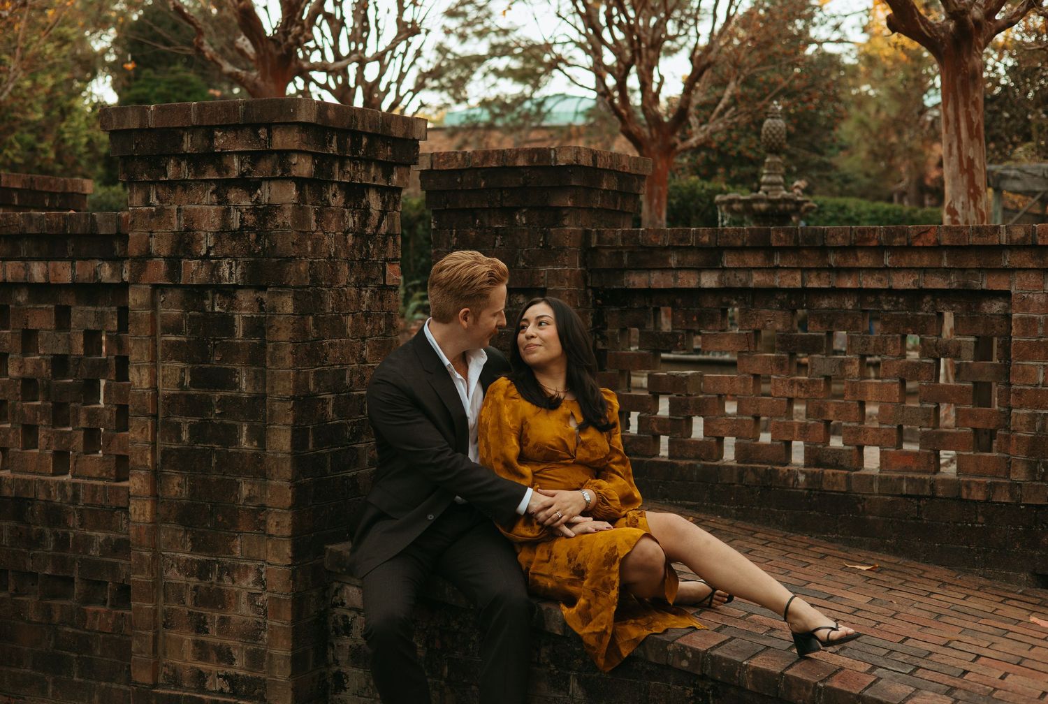 Couple sitting intimately on brick garden wall surrounded by trees in warm autumn light.
