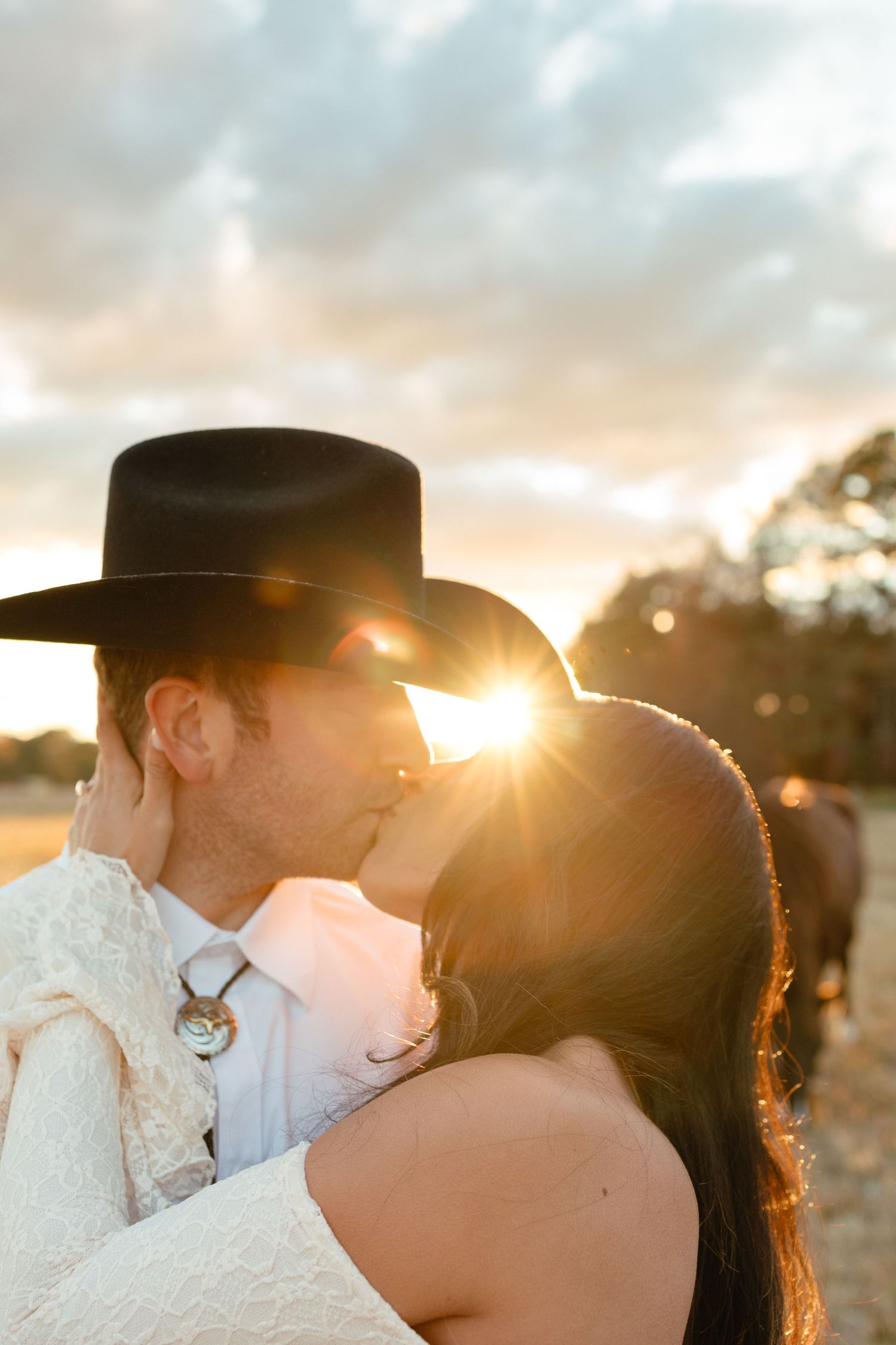 Couple shares romantic sunset kiss with groom in black cowboy hat at golden hour outdoor wedding.