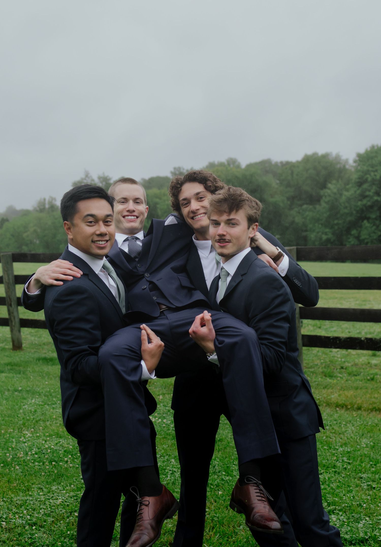 Four groomsmen in navy suits pose playfully outdoors, lifting one man on a green farm lawn.