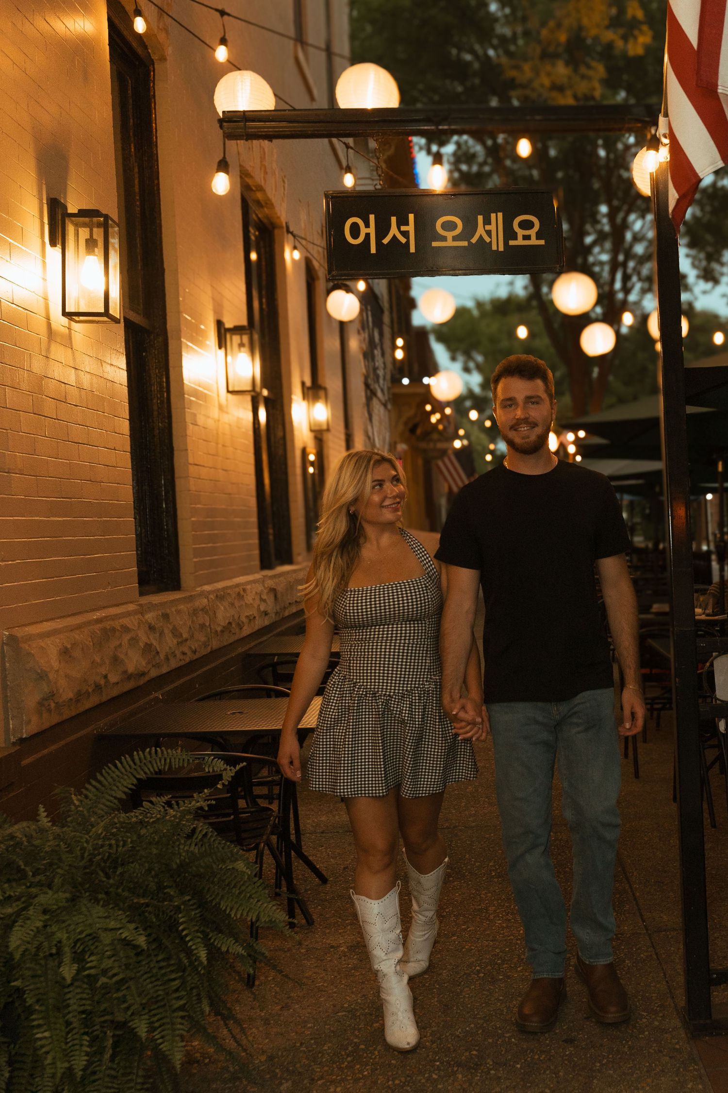 Couple holding hands walking past a Korean restaurant with string lights and neon sign at dusk.