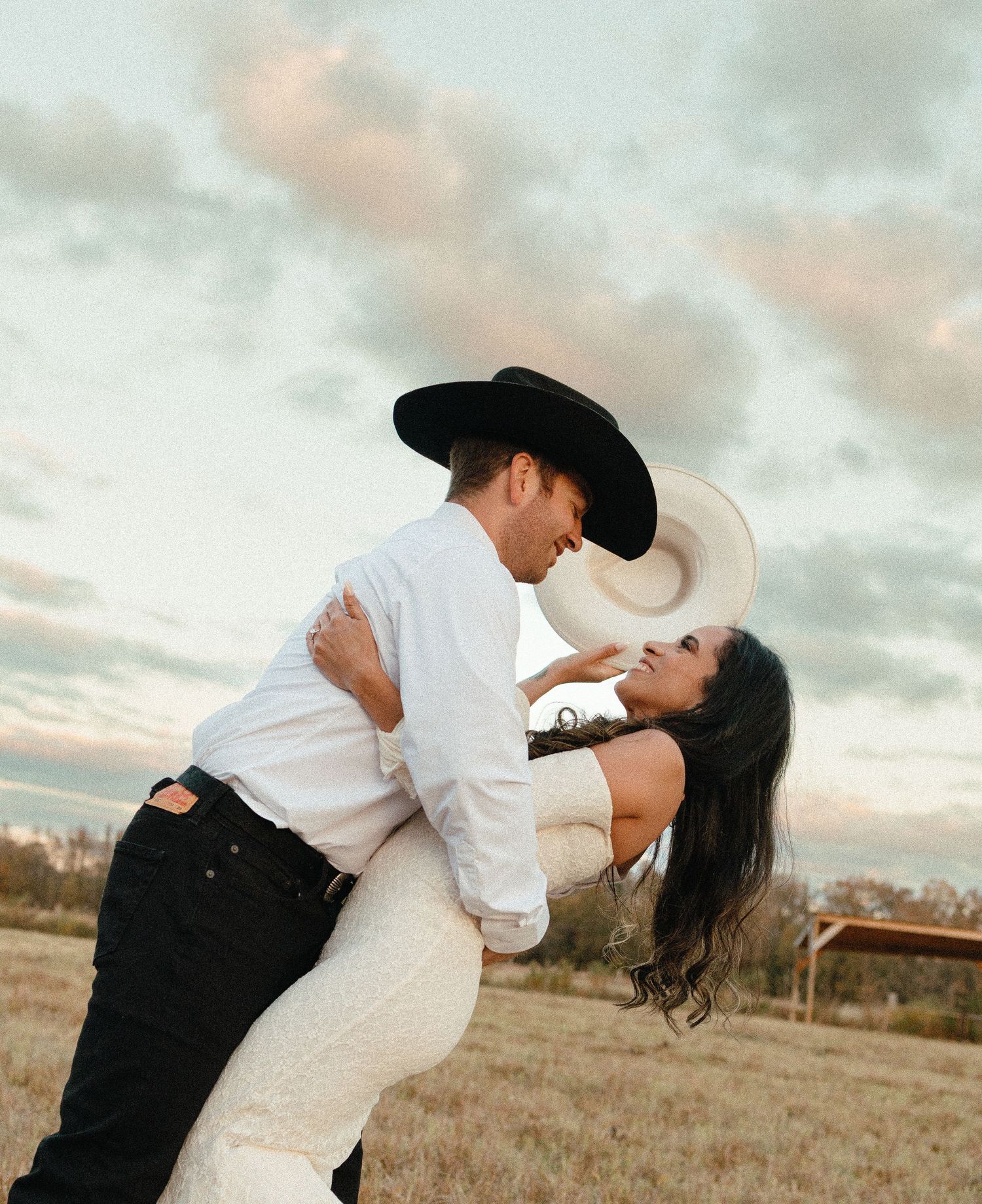 Groom in black cowboy hat dips bride in white dress during romantic outdoor engagement photo.