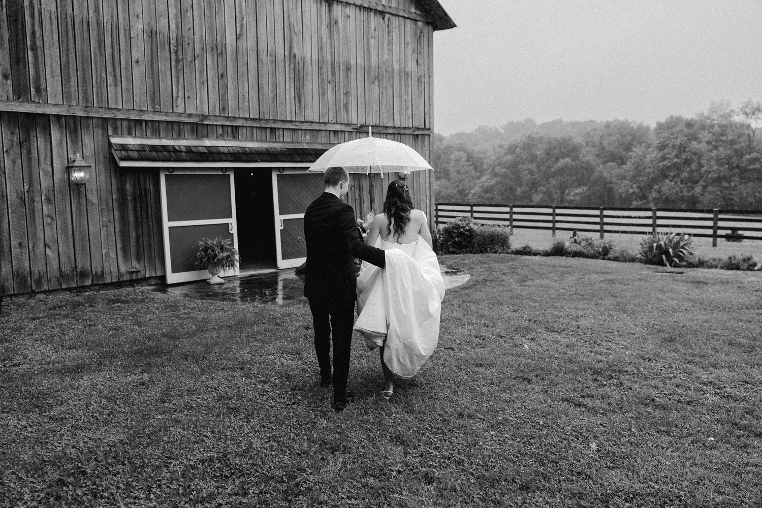 Black and white photo of newlyweds walking hand in hand under umbrella toward rustic barn in rain.