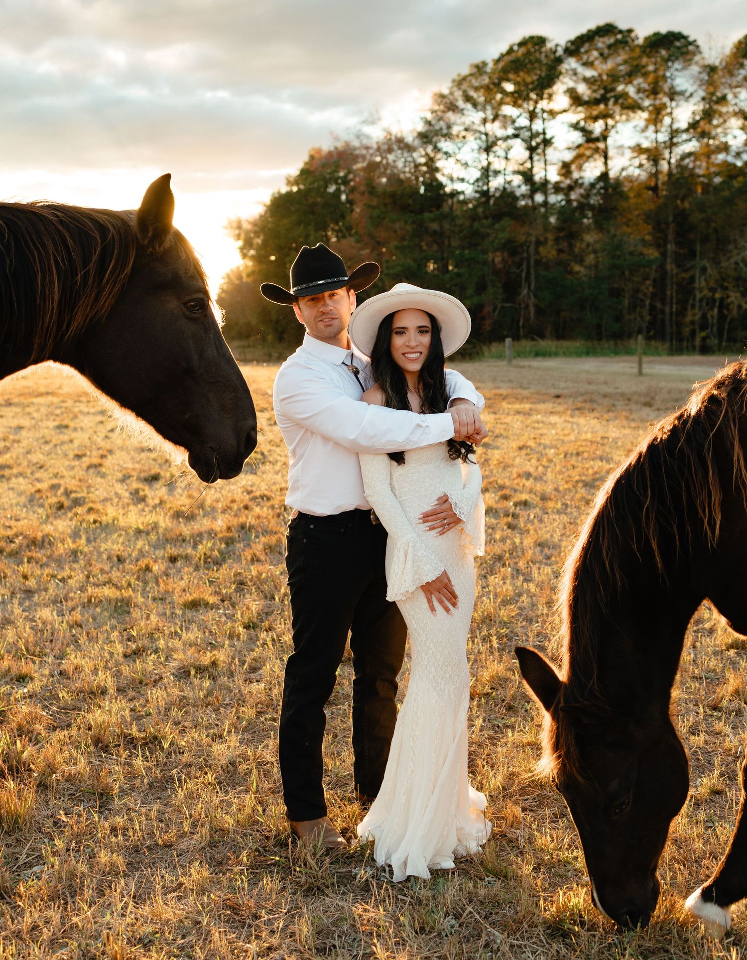 Couple in cowboy hats poses in golden field at sunset surrounded by dark horses in wedding attire.
