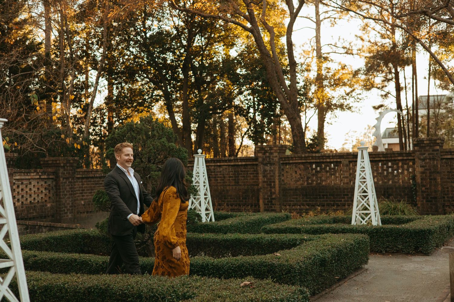 Man on one knee proposing to woman in mustard dress in elegant formal garden setting.