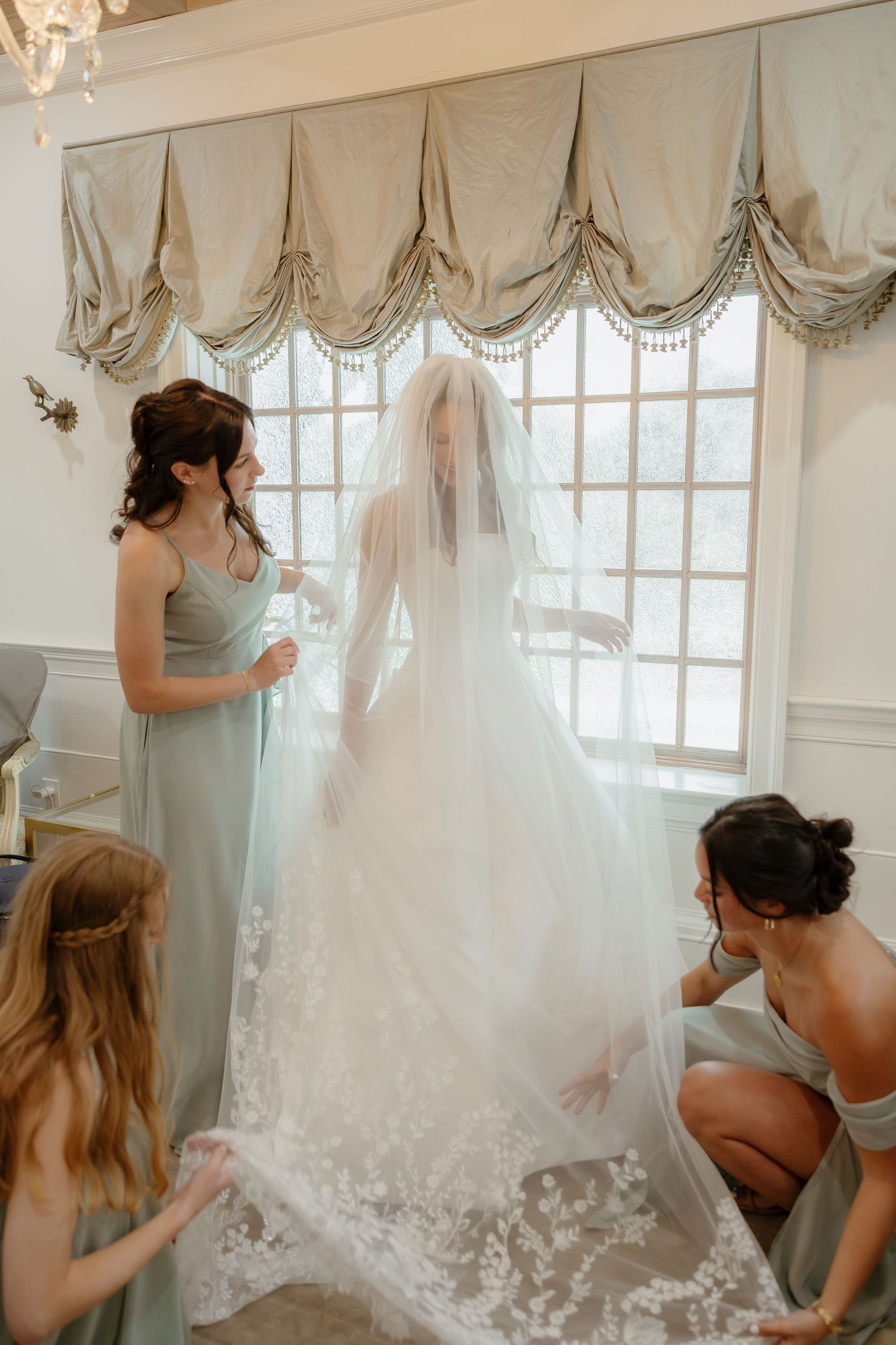 Bridesmaids in sage green dresses help bride arrange lace wedding gown and veil near elegant window.