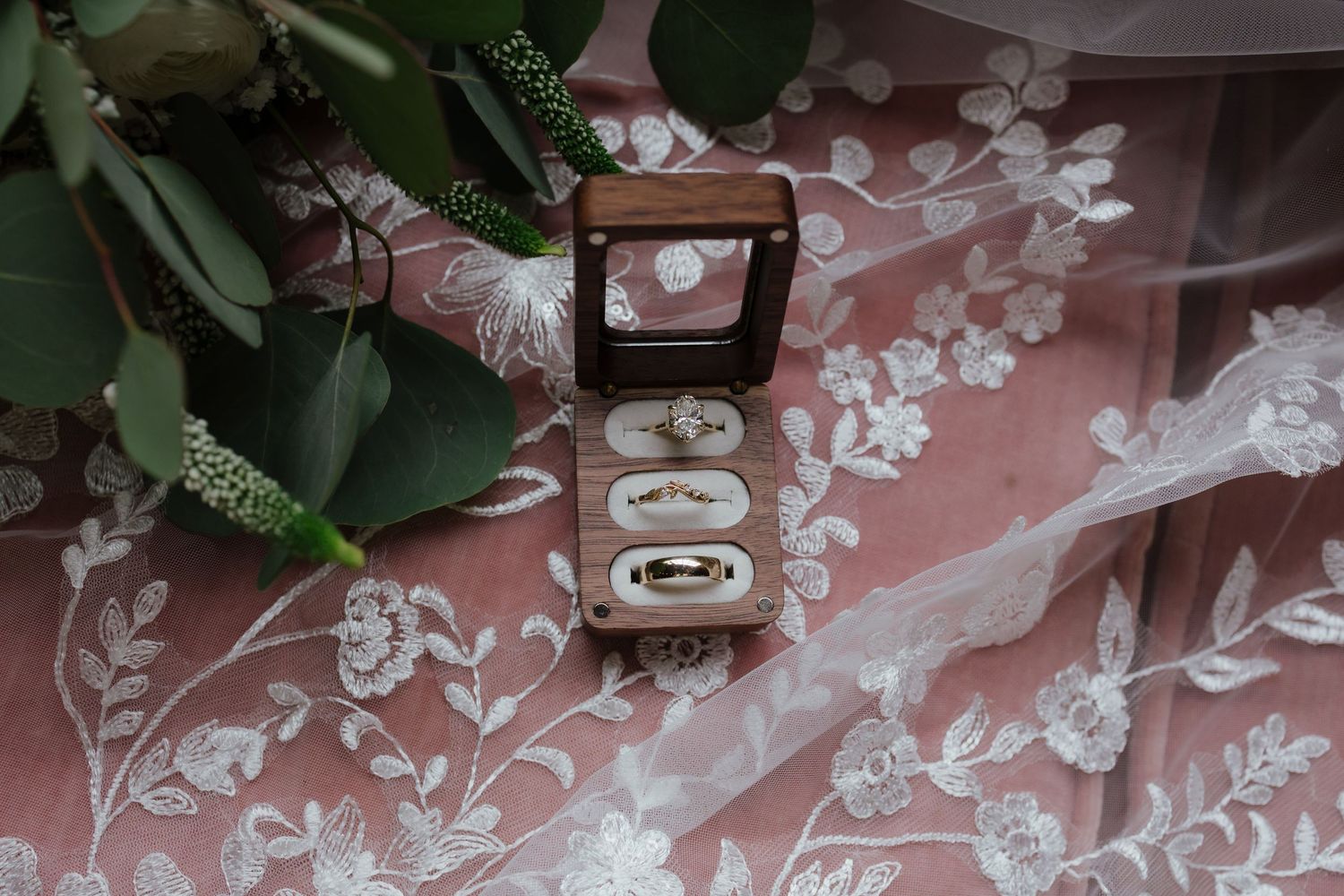 Wedding rings displayed in a wooden ring box on floral lace veil with greenery bouquet.