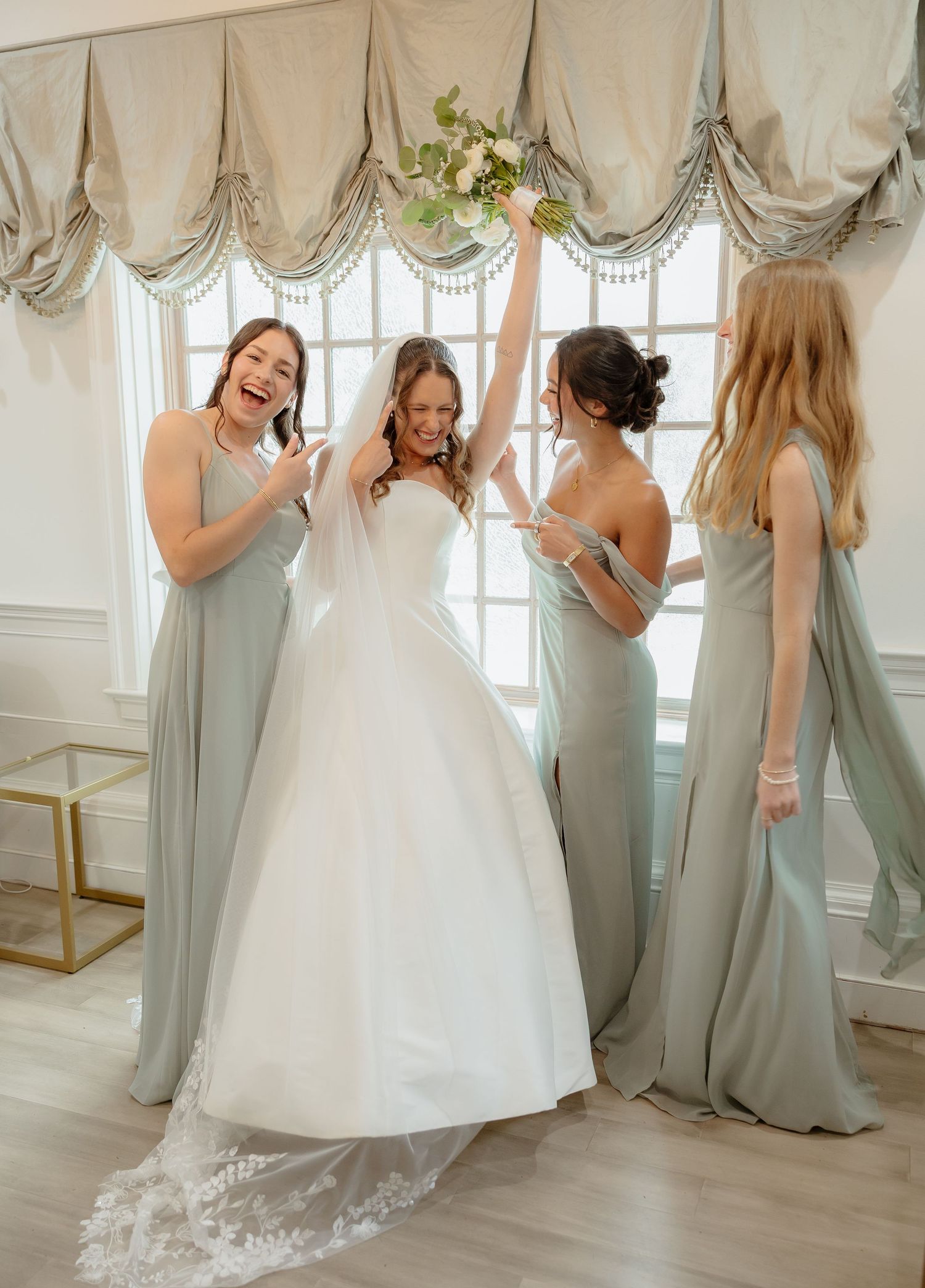 Bride in white ball gown celebrates with three bridesmaids in sage green dresses near bright window.