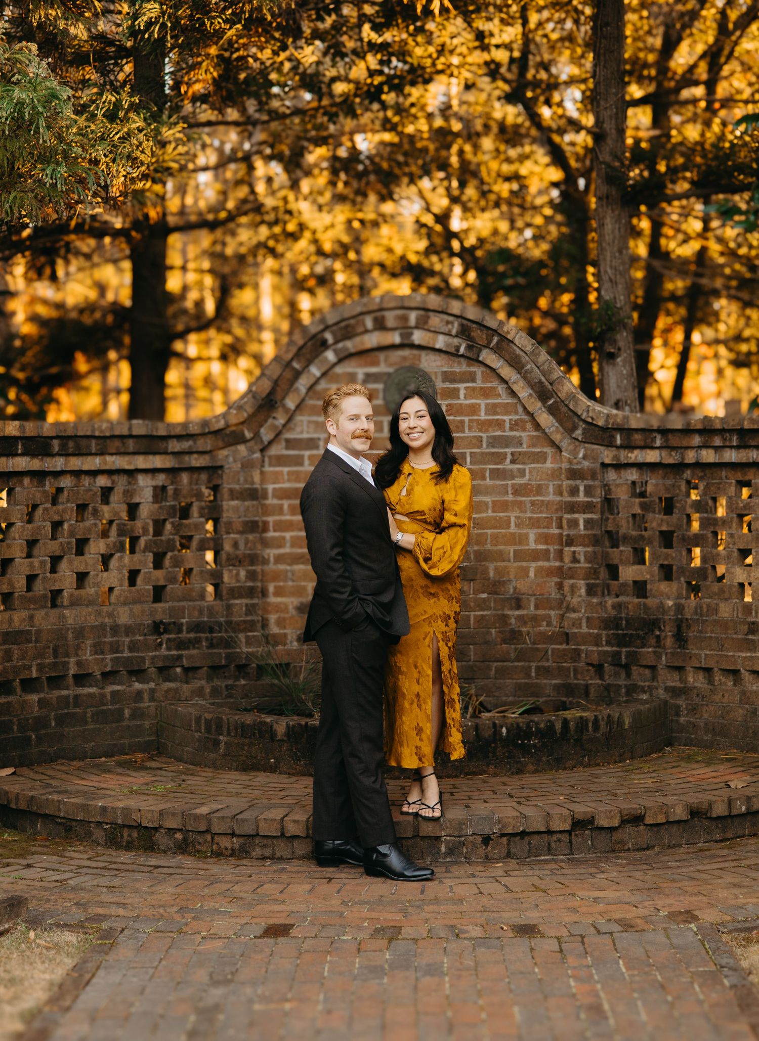 Couple posing together in front of ornate brick lattice wall surrounded by golden autumn foliage.