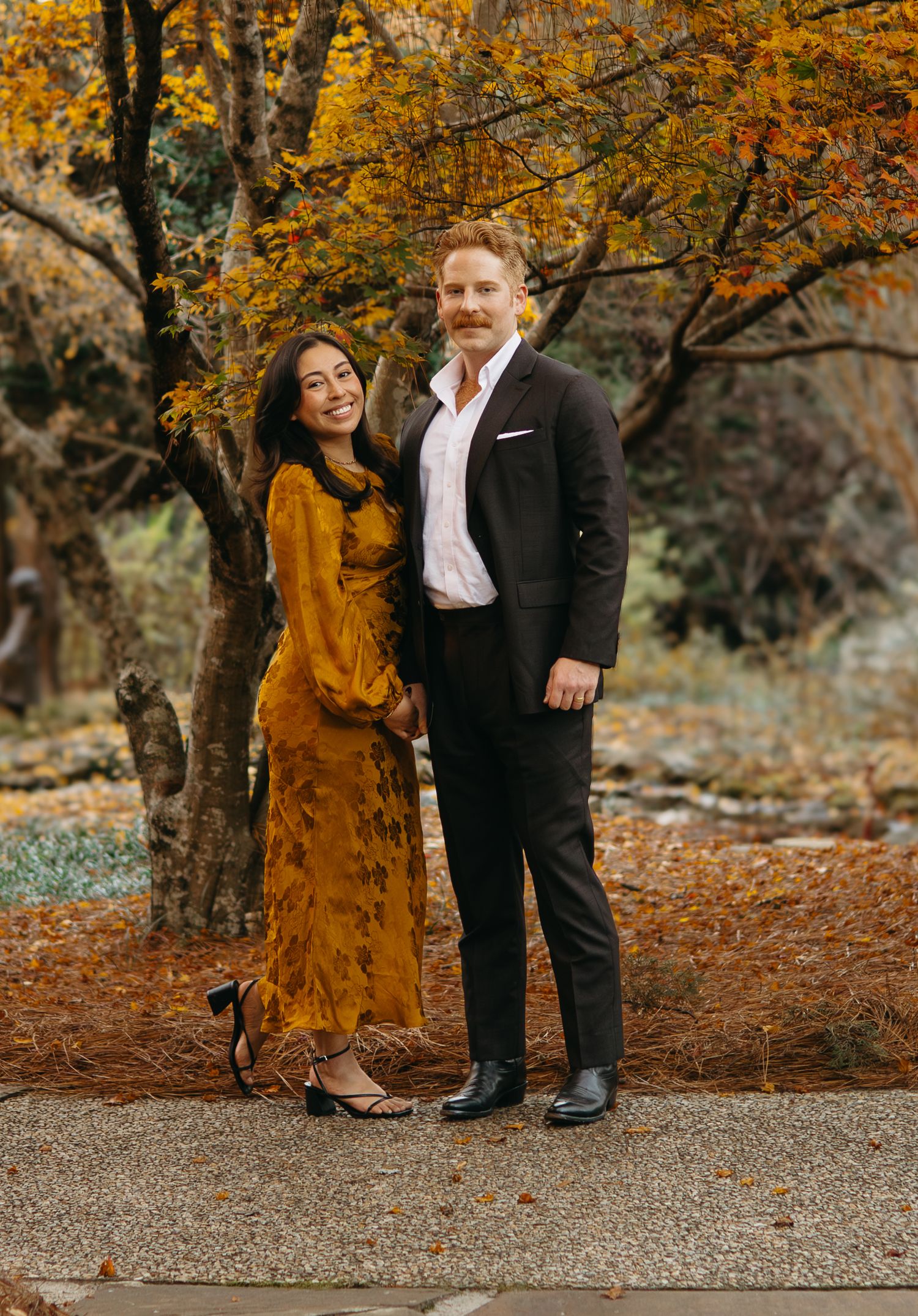 Smiling couple posing together in a park with vibrant fall foliage in mustard and autumn tones.