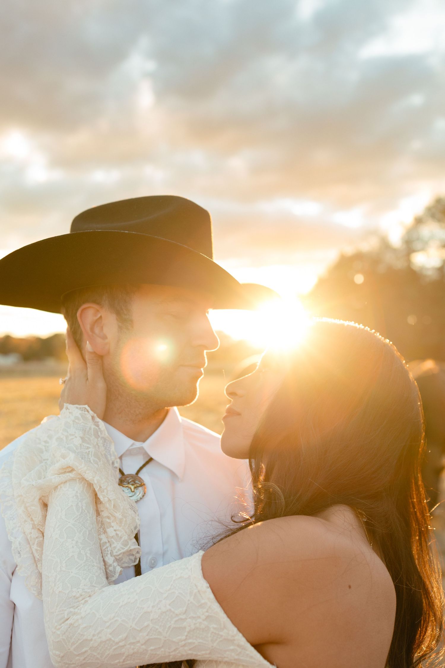Bride and groom nearly kiss bathed in a brilliant golden sunset lens flare in an open field wearing western attire.