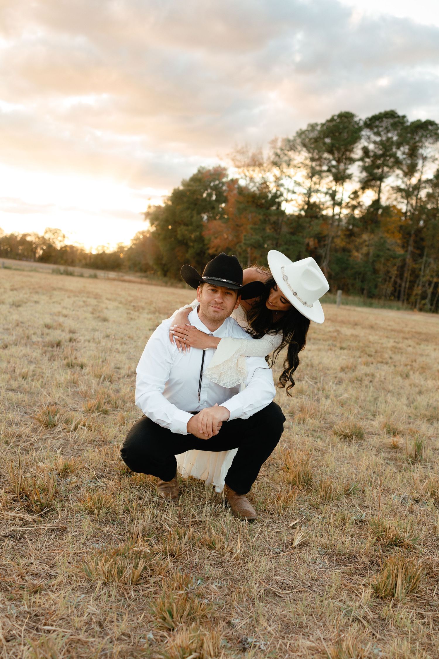 Couple in cowboy hats posing at golden hour in a dry field, man crouching while woman embraces him.