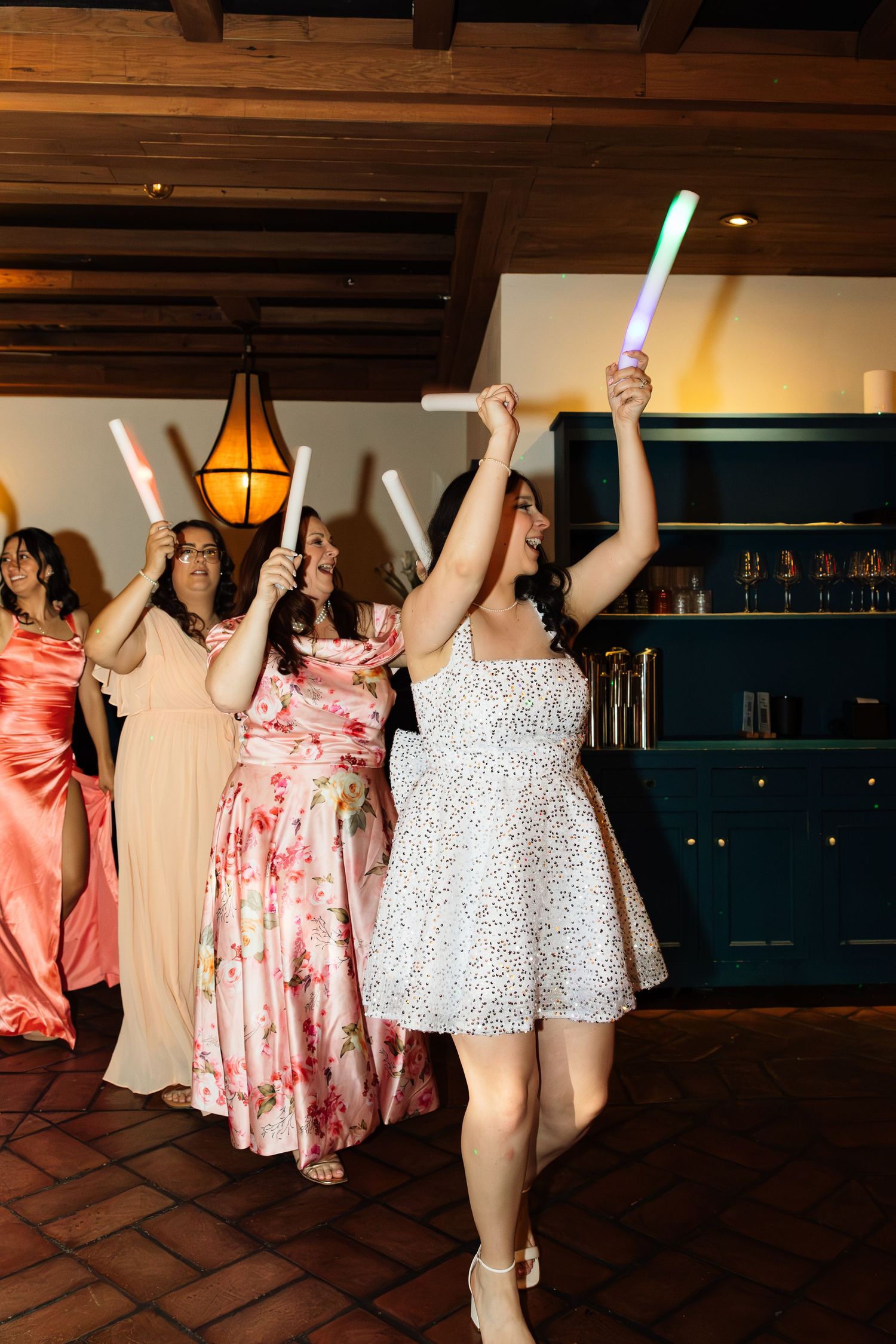 Bridesmaids dancing and waving glow sticks at a lively indoor wedding reception celebration.