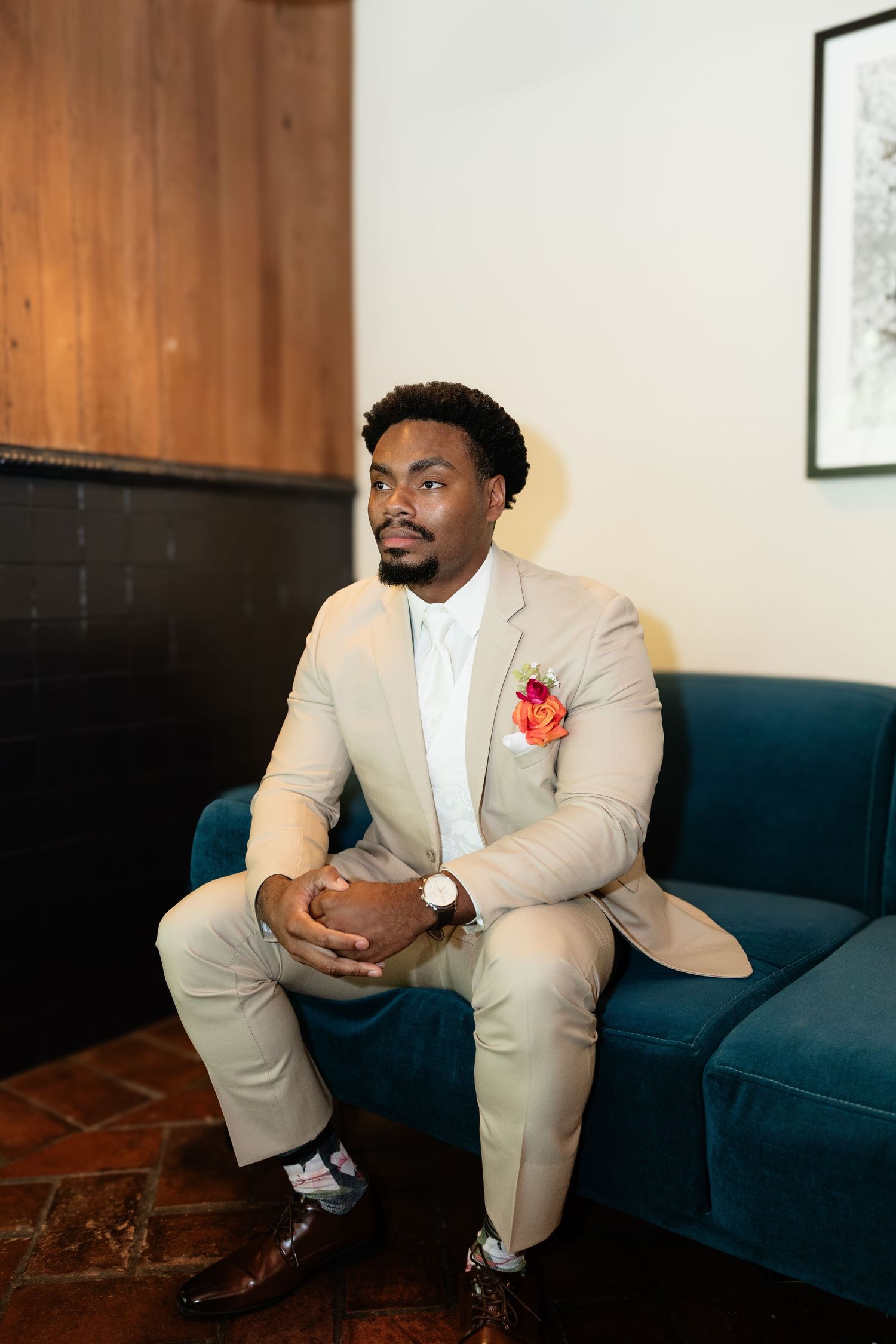 Groom in tan suit with colorful pocket square sitting on teal velvet sofa looking away thoughtfully.