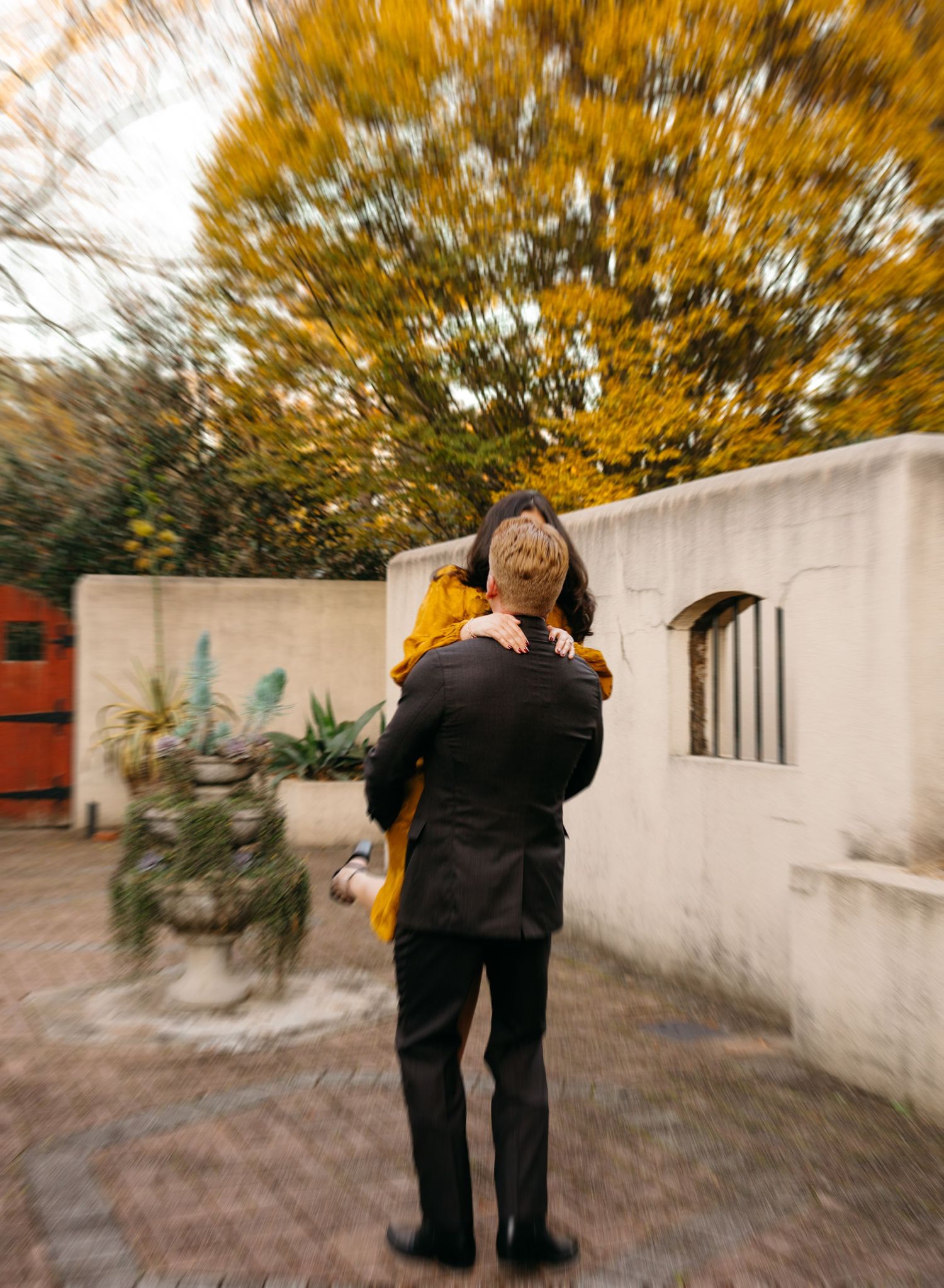 Man in black suit lifts woman in mustard dress, embracing her against a backdrop of golden autumn trees.