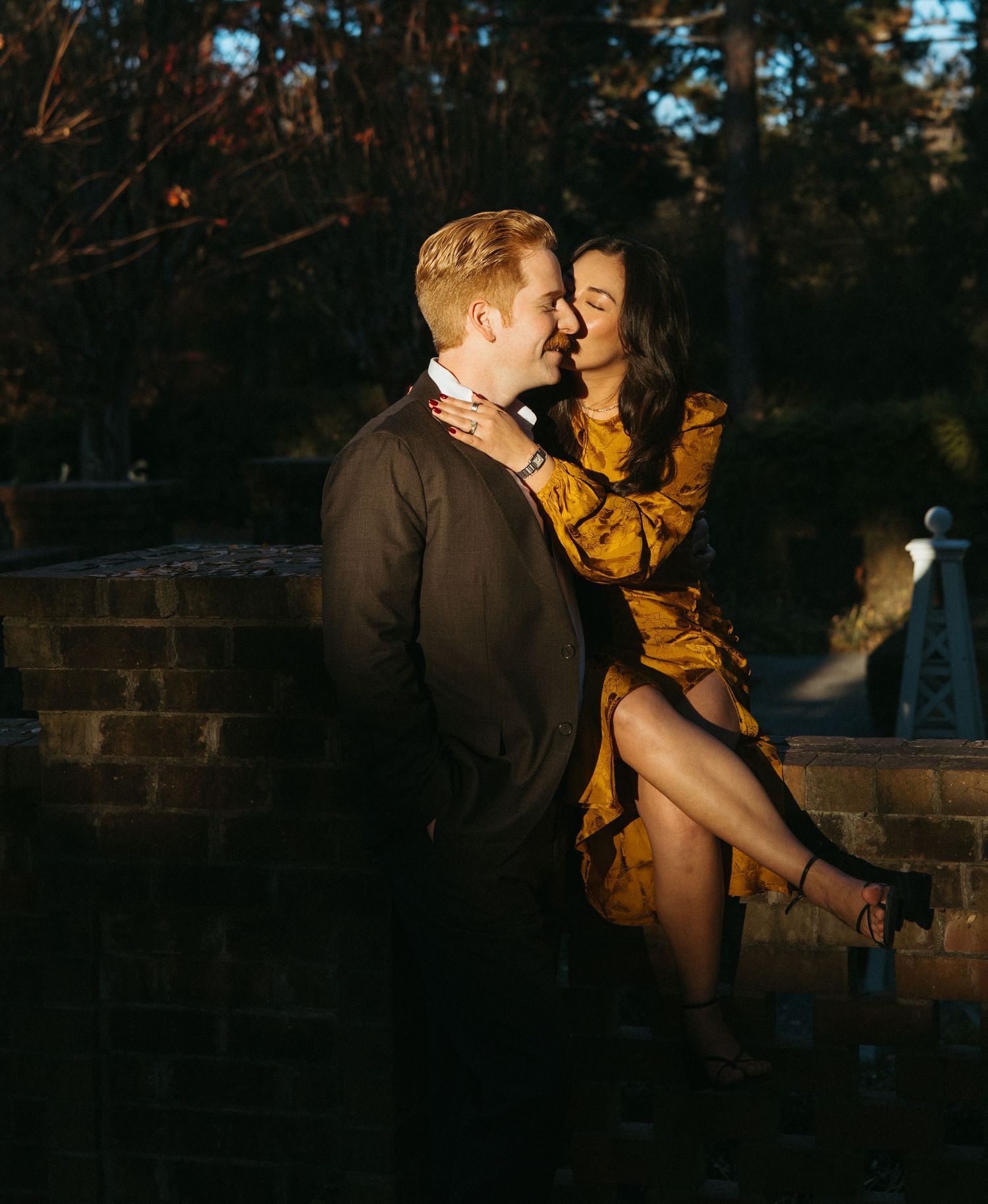 Couple sharing an intimate moment on a brick wall at golden hour in a garden.