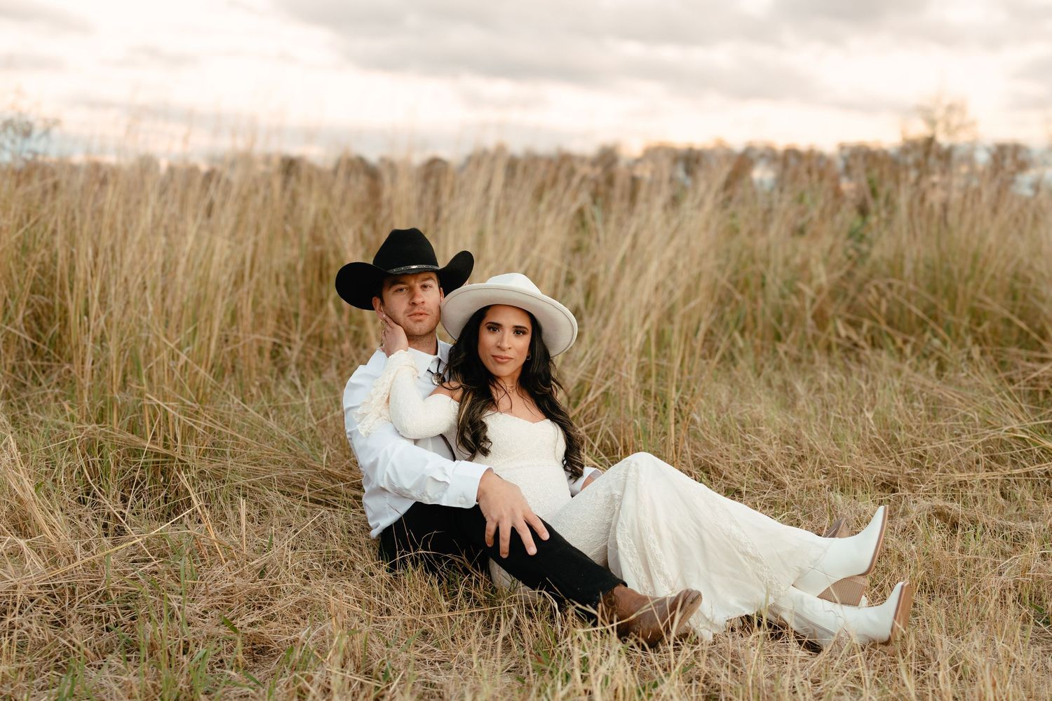 Couple in western wedding attire with cowboy hats posing in tall dry grass field at sunset.