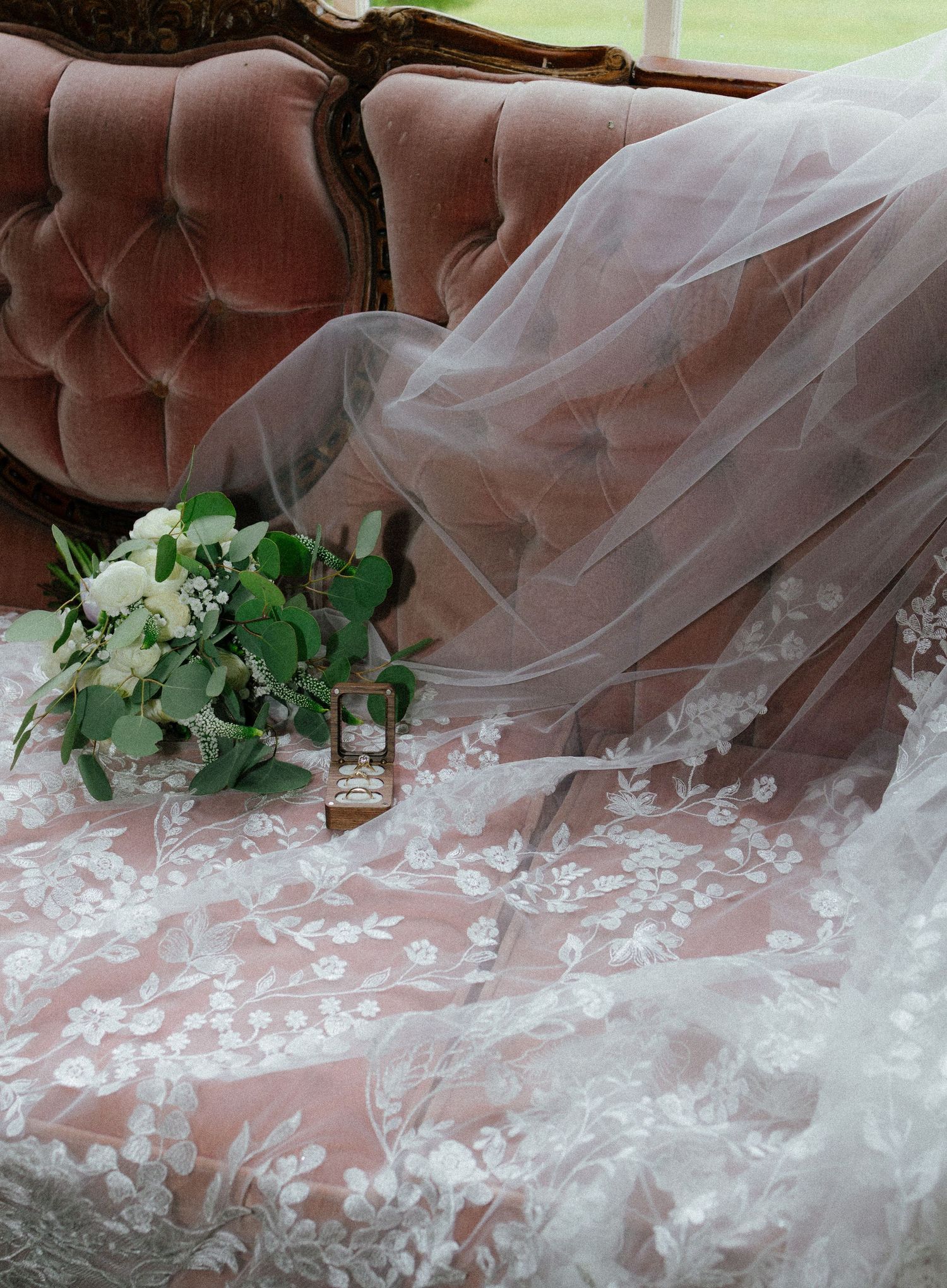 Close-up of intricate lace wedding gown and veil draped over pink velvet sofa with bridal bouquet.