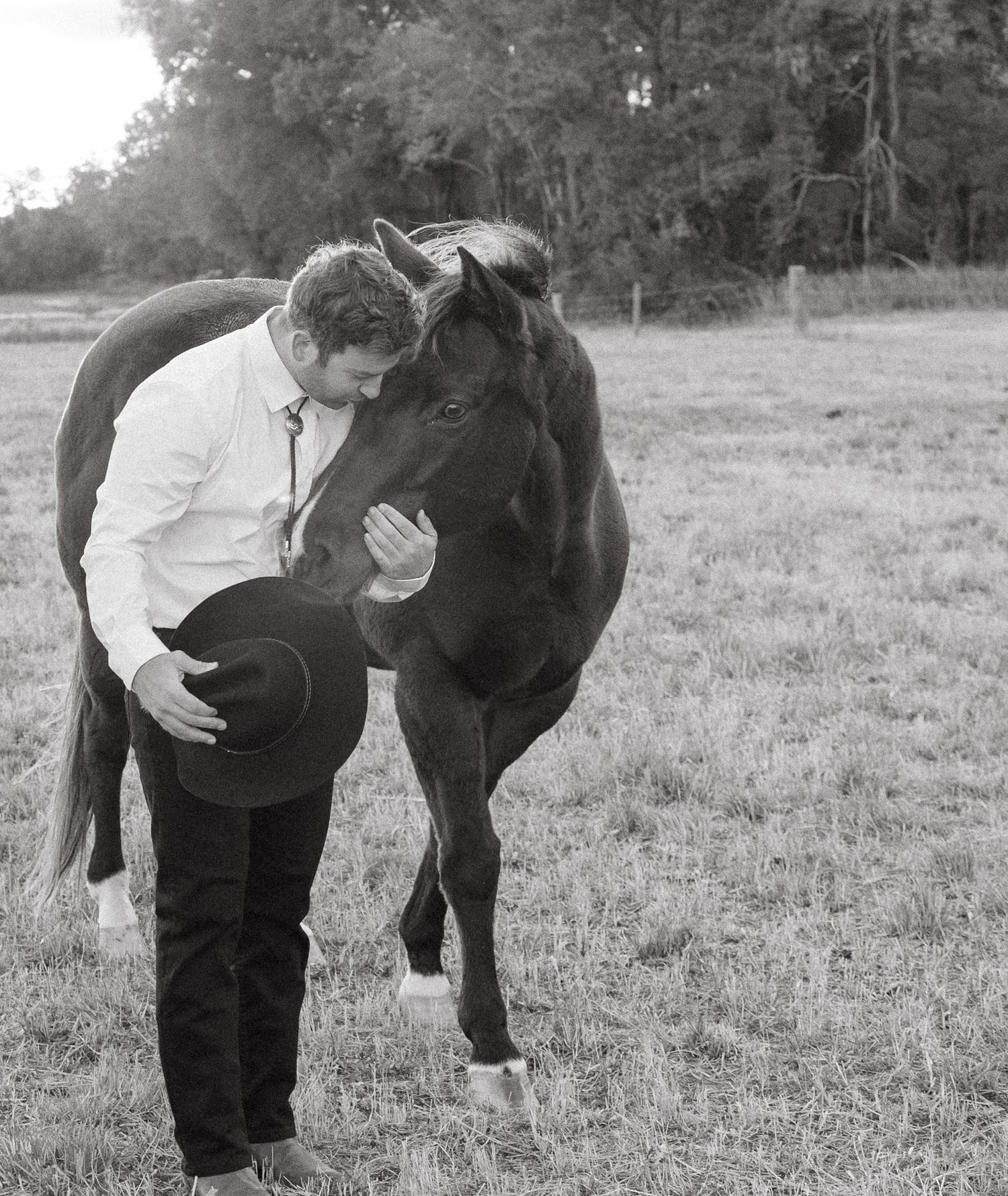 Black and white photo of a man in a white shirt kissing a dark horse in an open field.