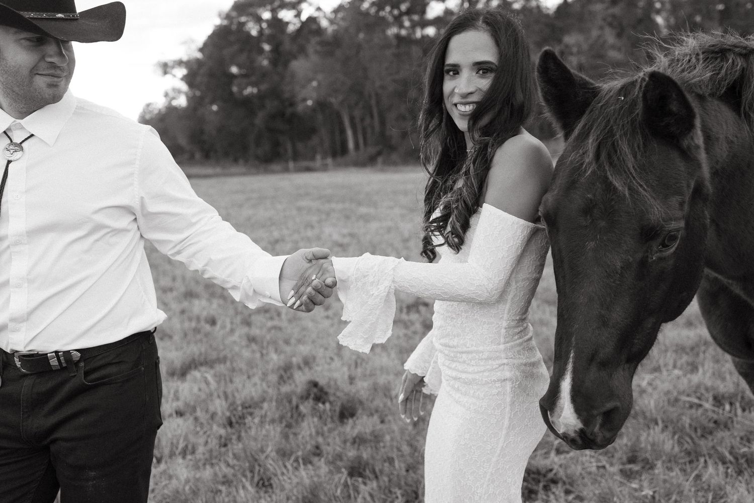 Black and white photo of smiling bride in fitted gown holding groom's hand beside a horse in an open field.