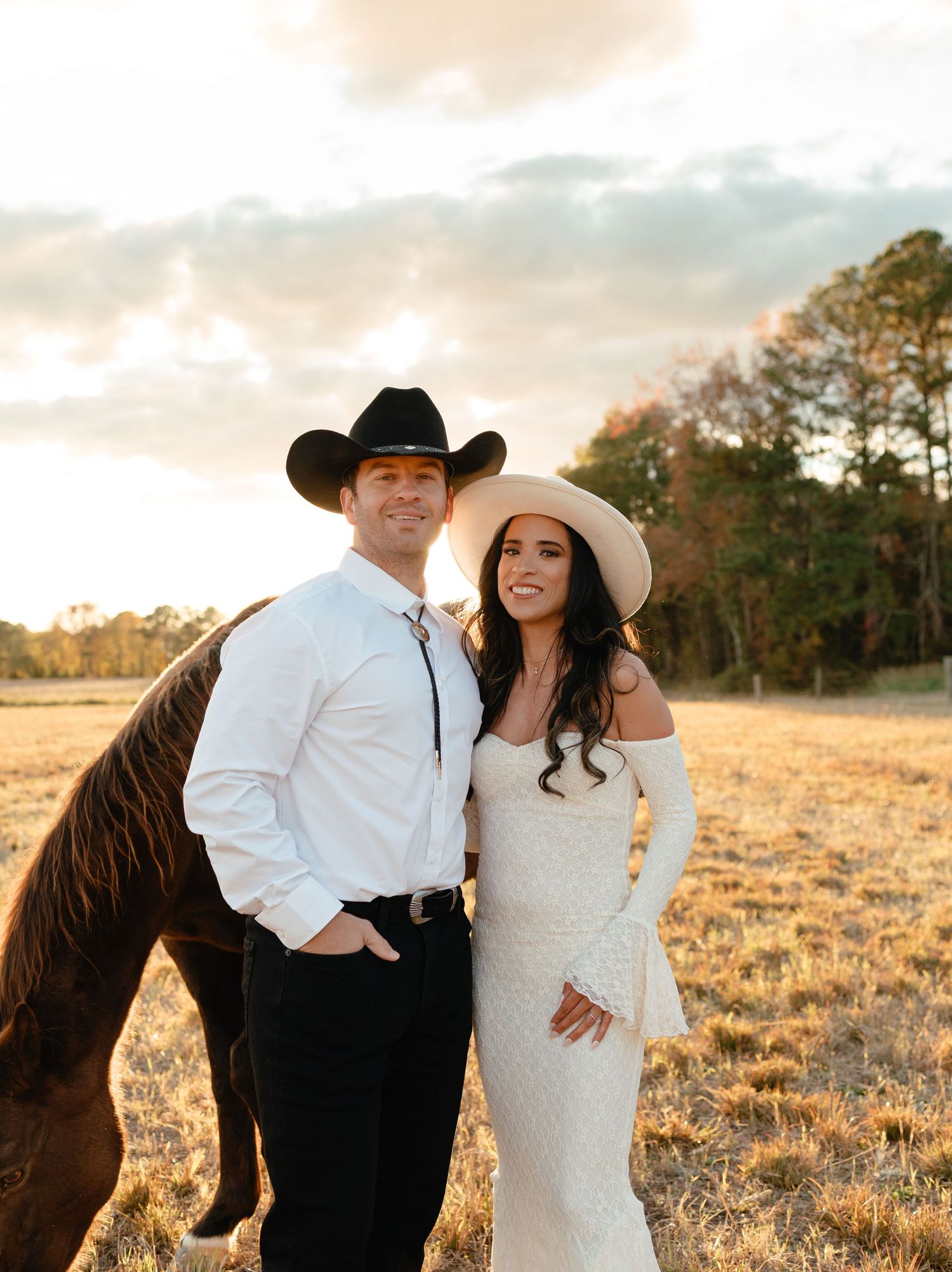 Couple in coordinating cowboy hats posing with a chestnut horse in a golden field at sunset.