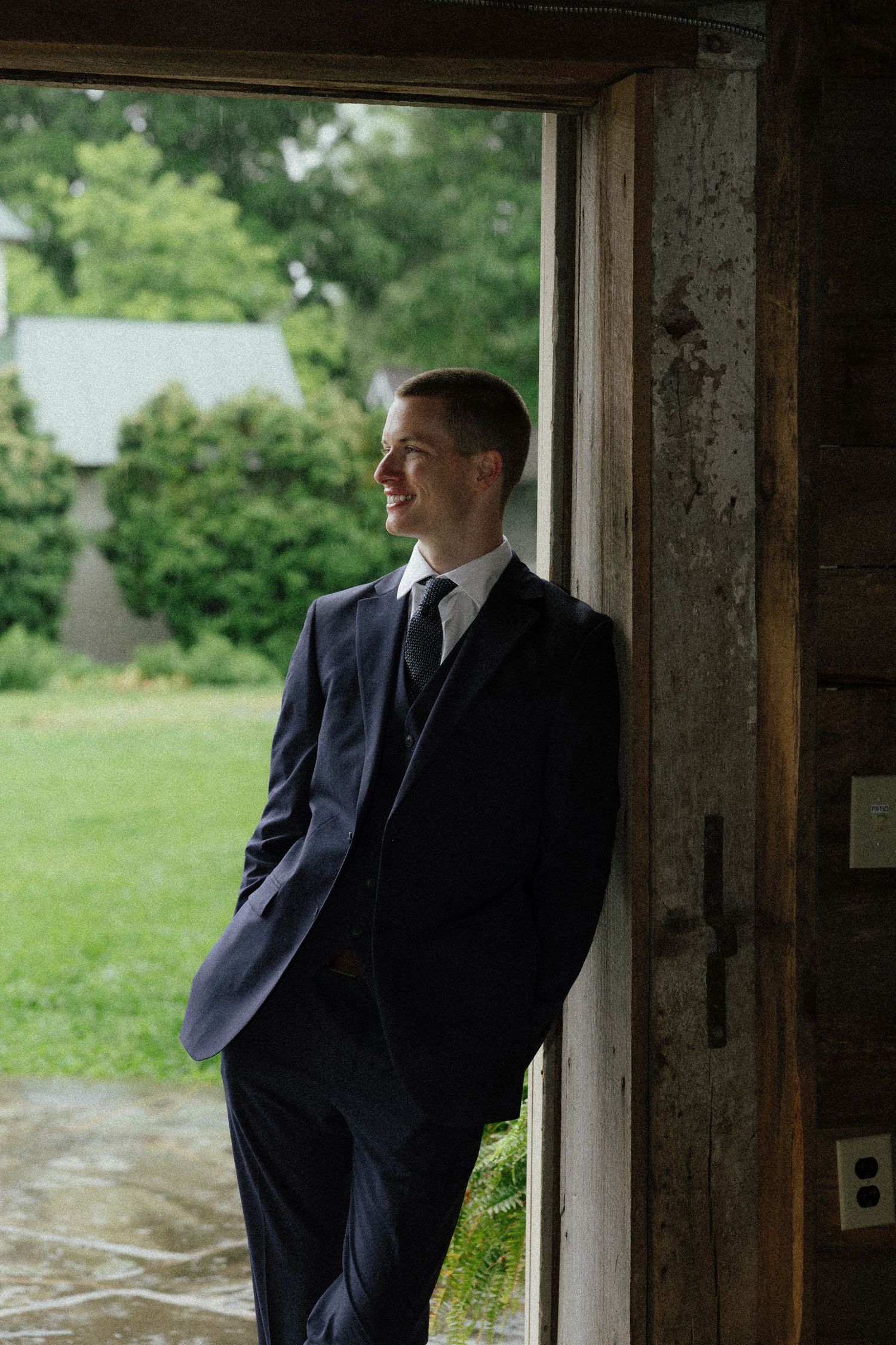 Dapper groom in navy suit leans against rustic barn doorway smiling at lush green outdoor setting.
