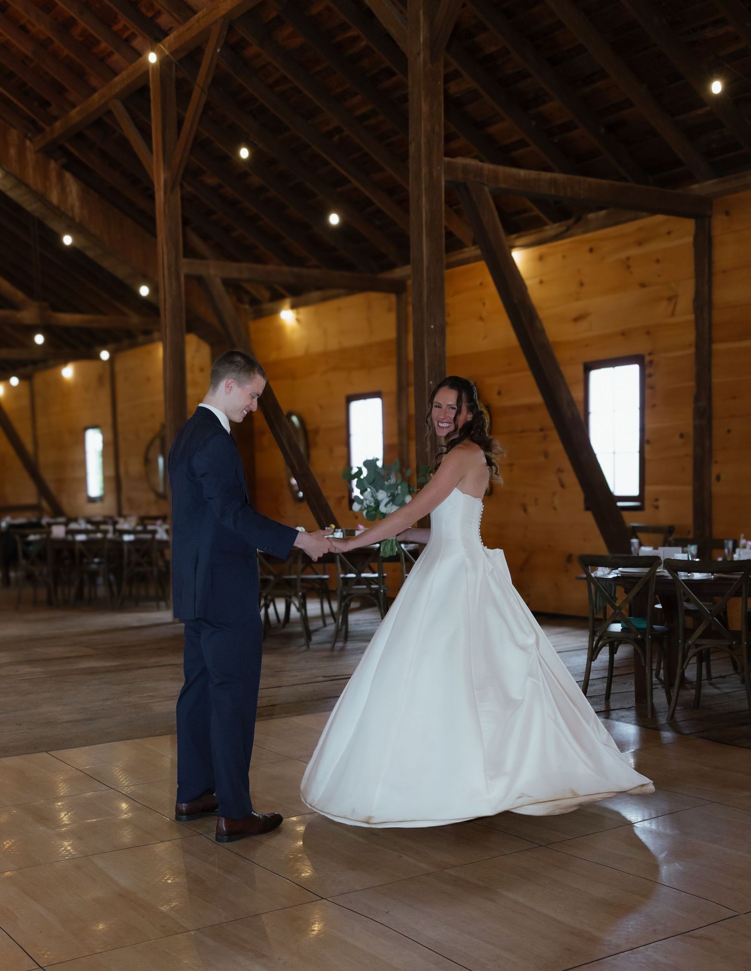Bride and groom share first dance on wooden floor inside rustic barn wedding venue with string lights.