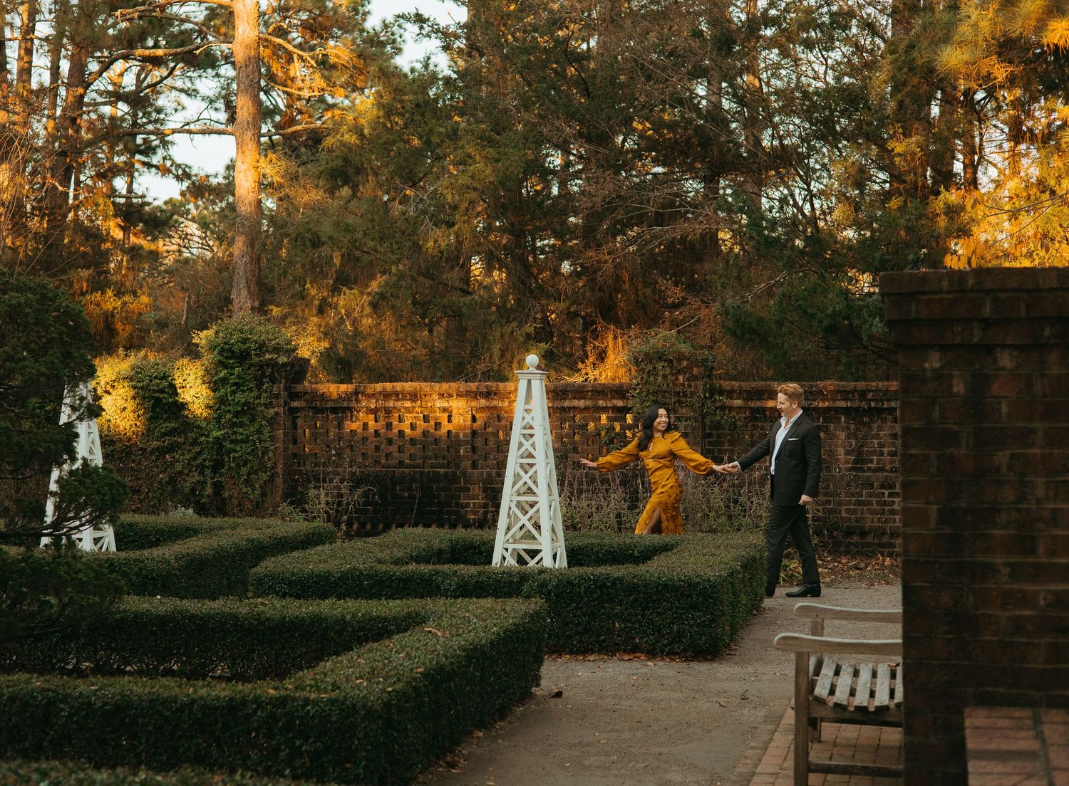 Couple holding hands walking through formal garden with boxwood hedges at golden hour sunset.
