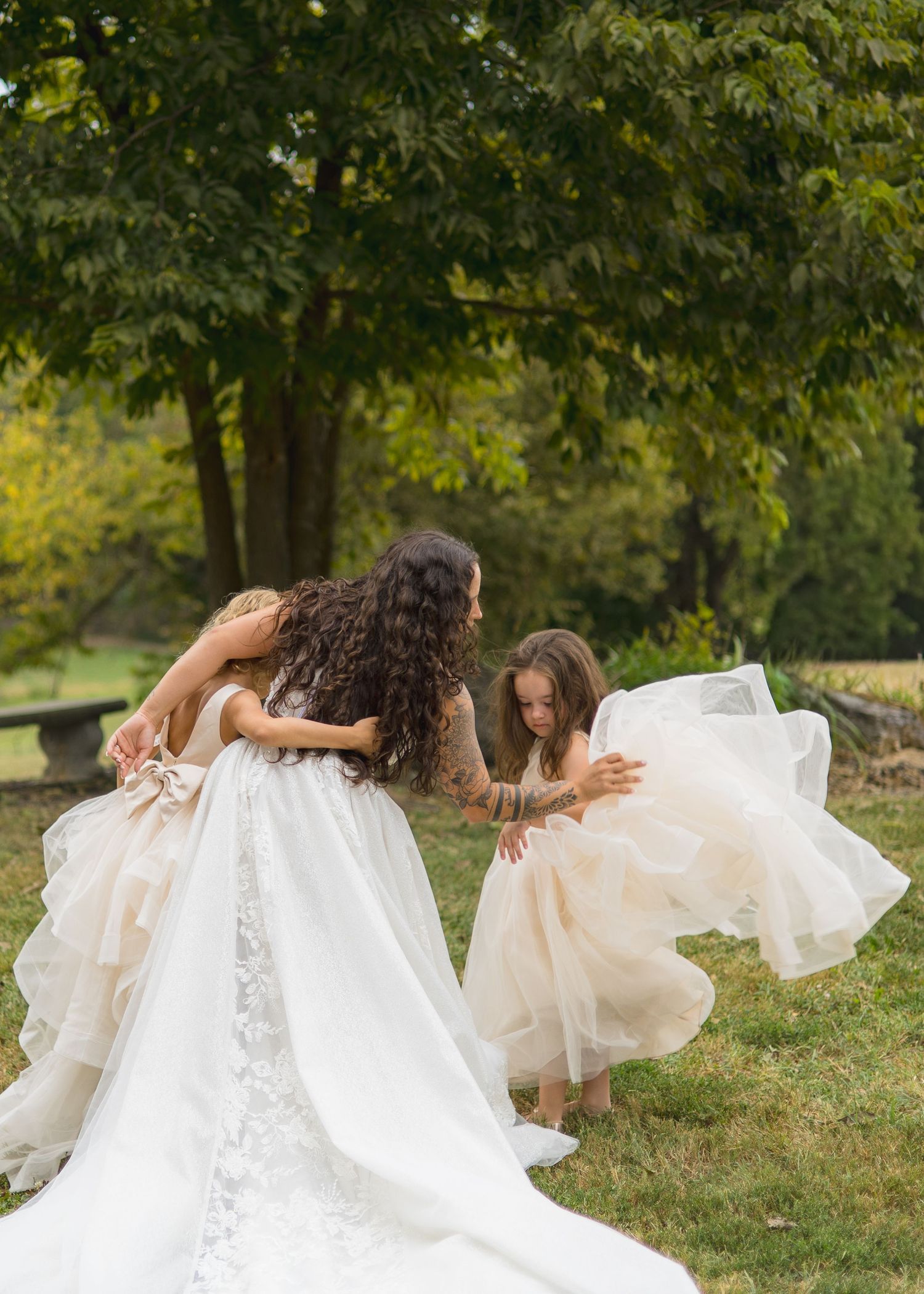 Tattooed bride in lace wedding gown twirls with two flower girls in champagne dresses outdoors.