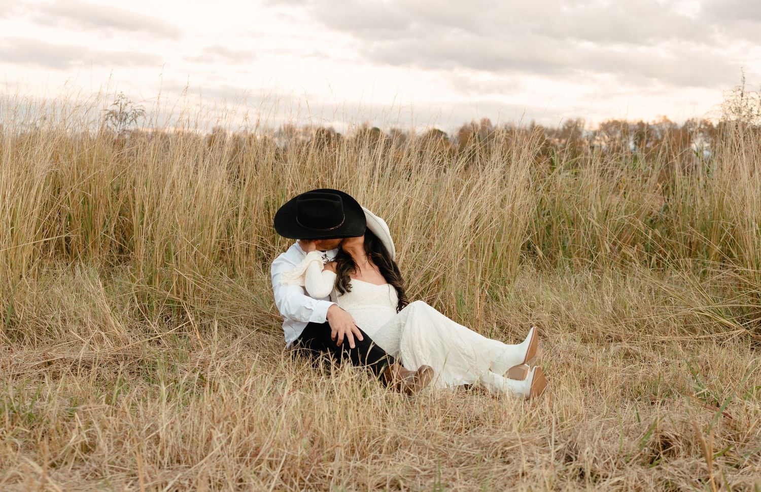 Bride and groom sharing a kiss while sitting in a rustic golden grass field at dusk.