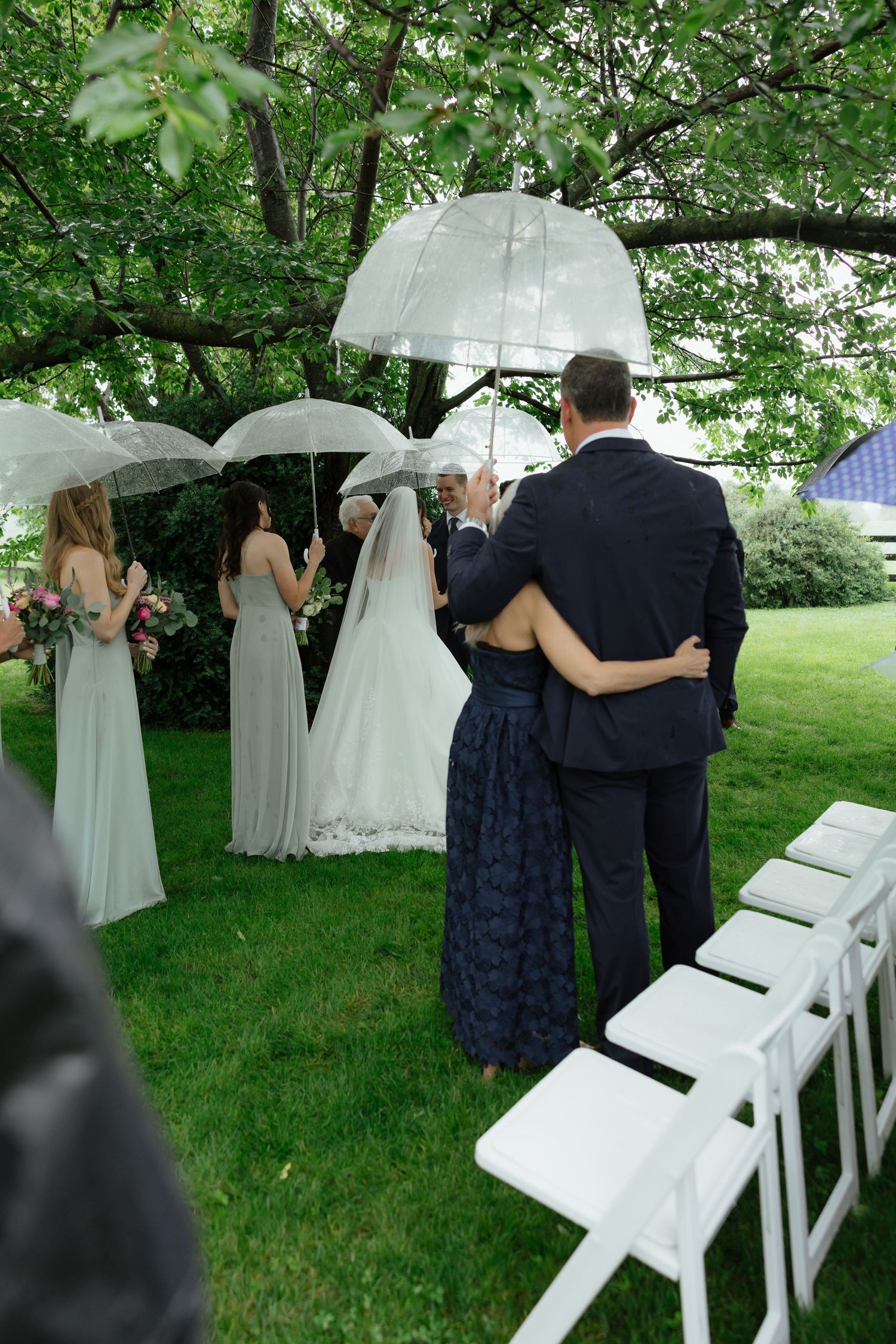 Wedding party with clear umbrellas during outdoor rainy ceremony as bride walks down the aisle.
