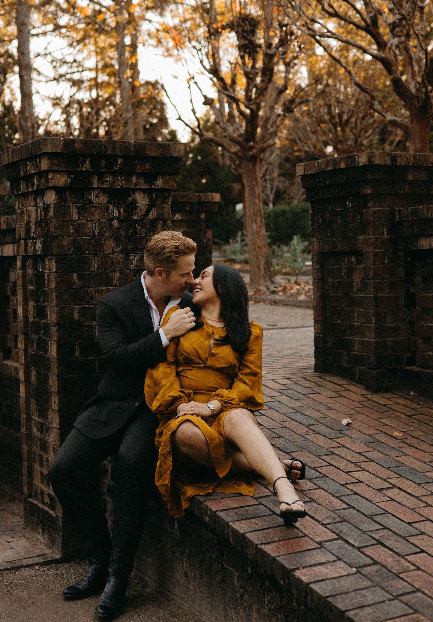 Couple sharing a tender moment on a brick ledge surrounded by autumn trees in warm evening light.