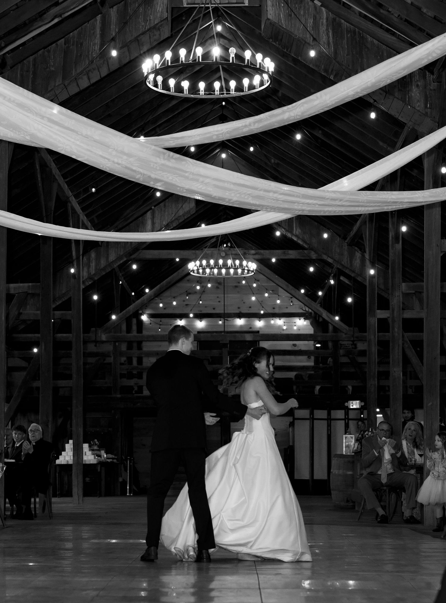Black and white photo of bride and groom dancing under chandeliers in a decorated barn.
