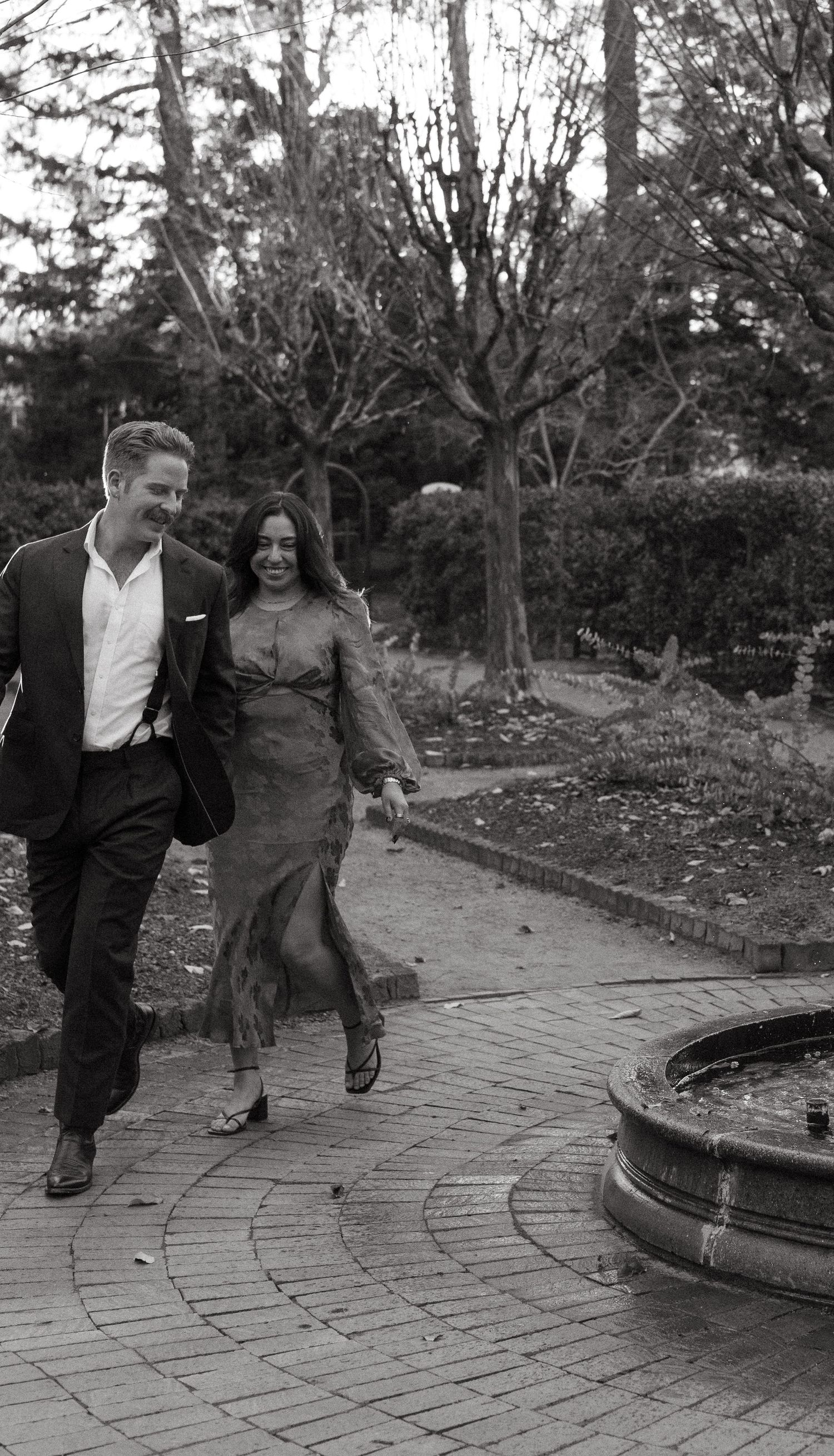 Black and white photo of a laughing couple walking hand in hand through an autumn garden path.
