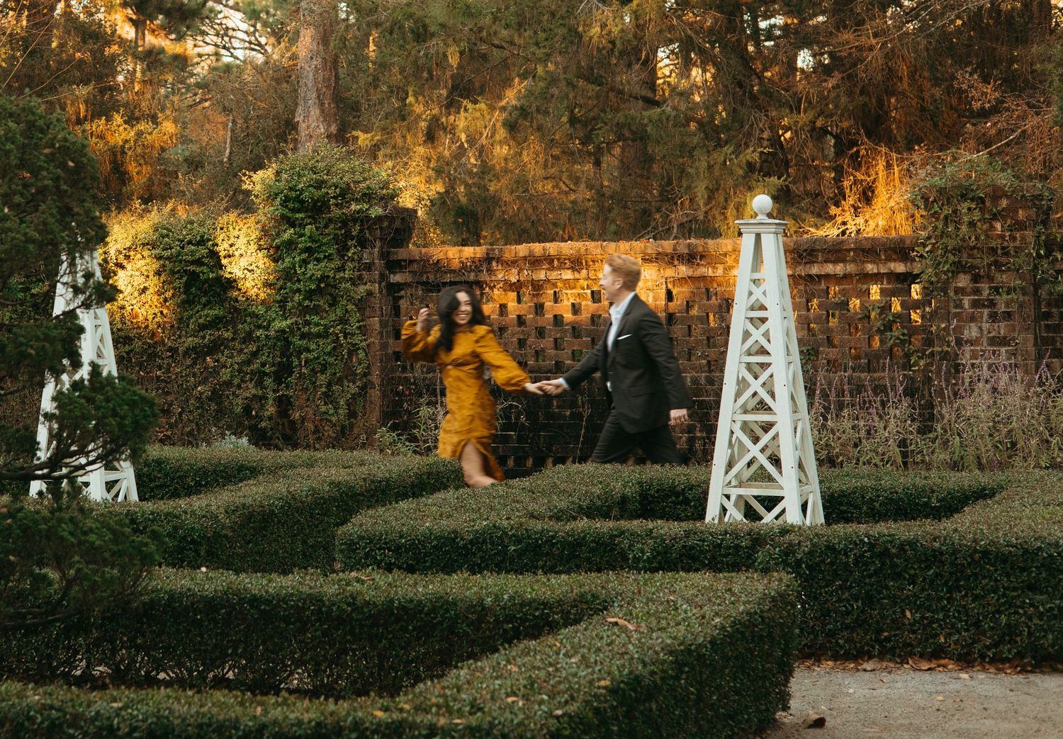 Couple holding hands and walking through a manicured garden maze at a warmly lit estate.