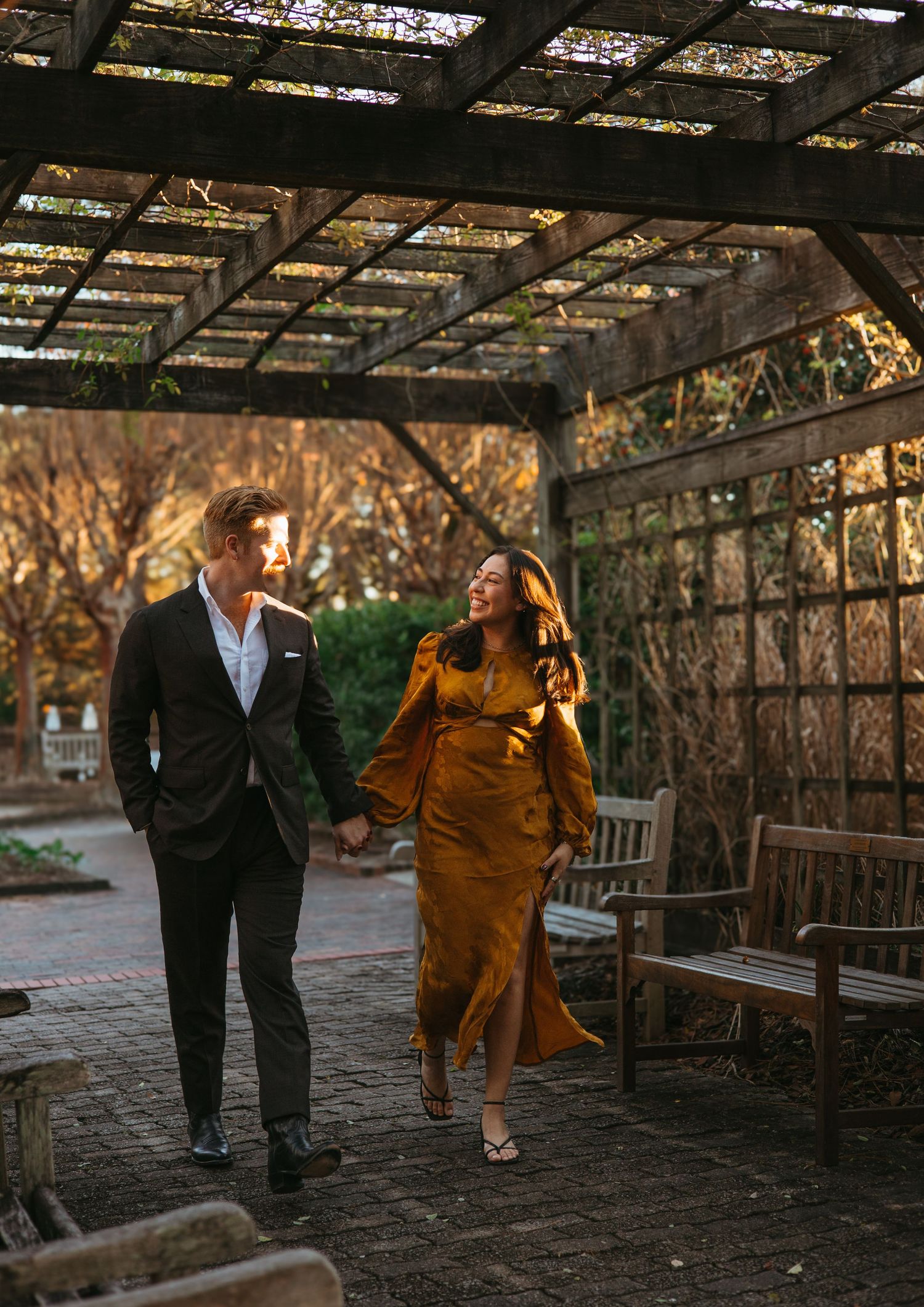 Couple walking hand in hand under a pergola, woman in golden dress, man in dark suit, garden setting at golden hour.