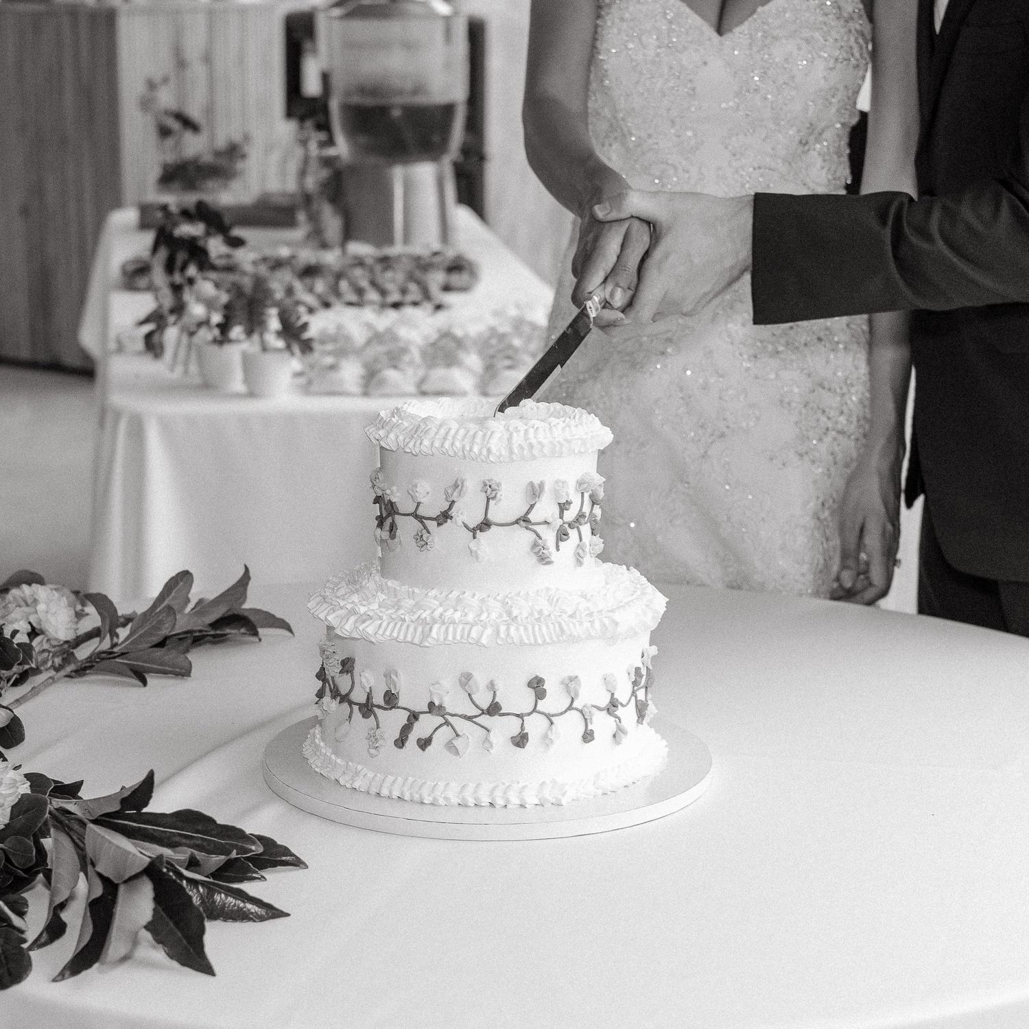 Bride and groom cutting a two-tier wedding cake decorated with floral vines in a black and white photo.