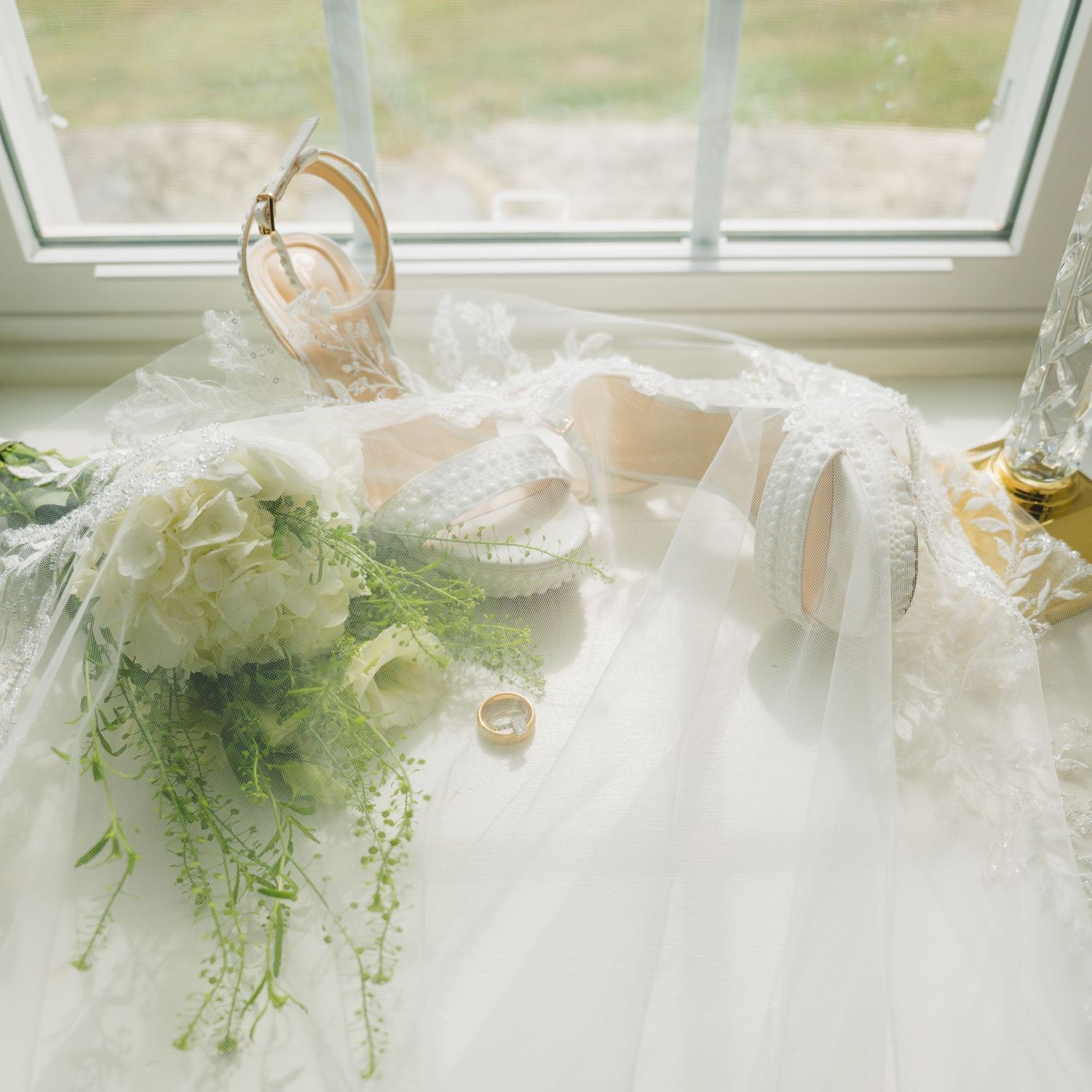A bridal flat lay featuring a white wedding gown, lace heels, bouquet, and gold rings by a window.