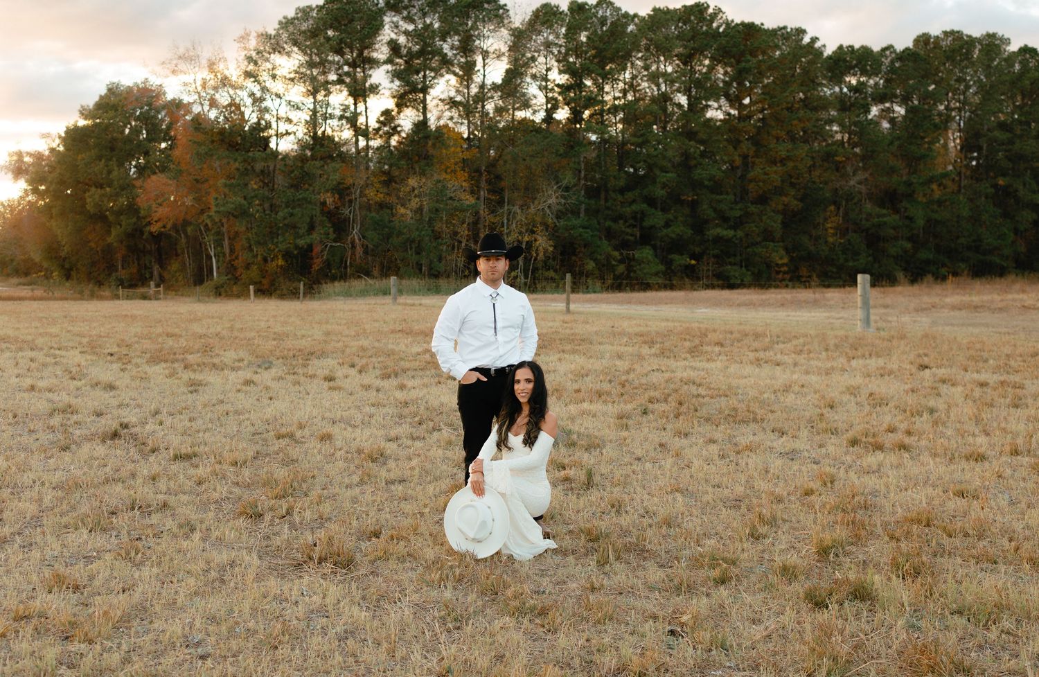 Couple in western attire posing in an open field at sunset, woman in white dress kneeling.
