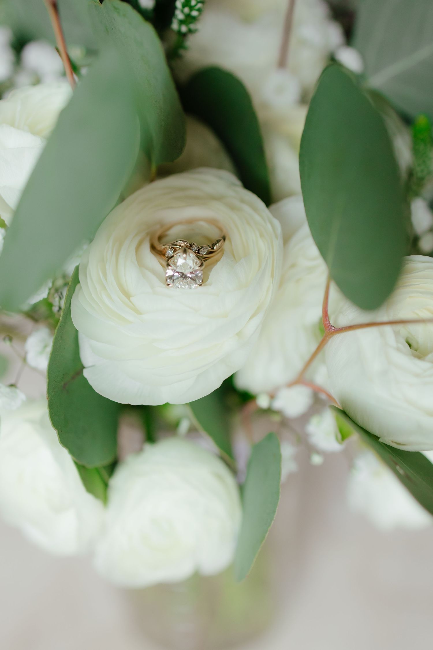 Gold diamond engagement ring nestled in white ranunculus flower surrounded by eucalyptus greenery.