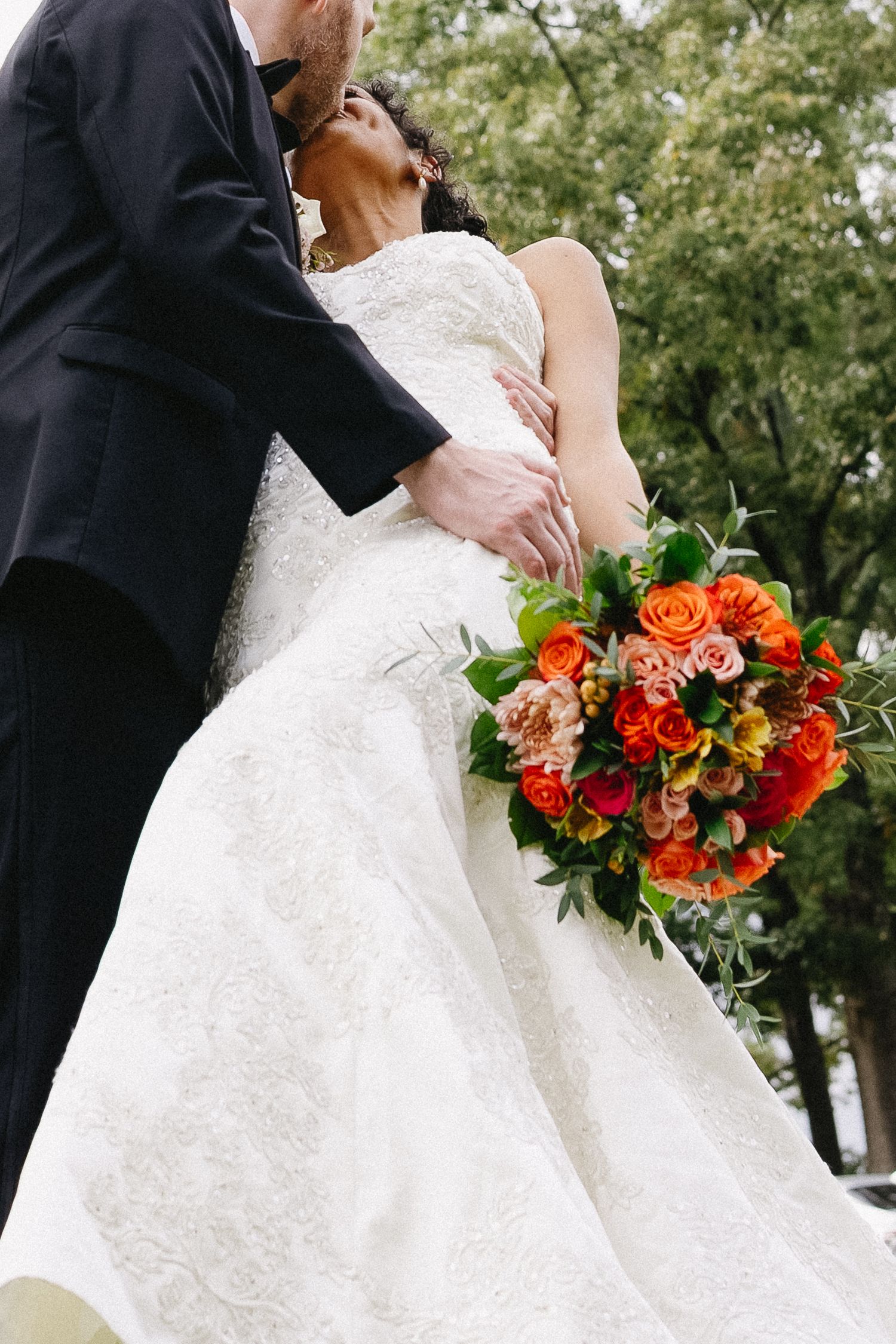 Bride and groom kissing outdoors with vibrant orange and red rose bridal bouquet in foreground.