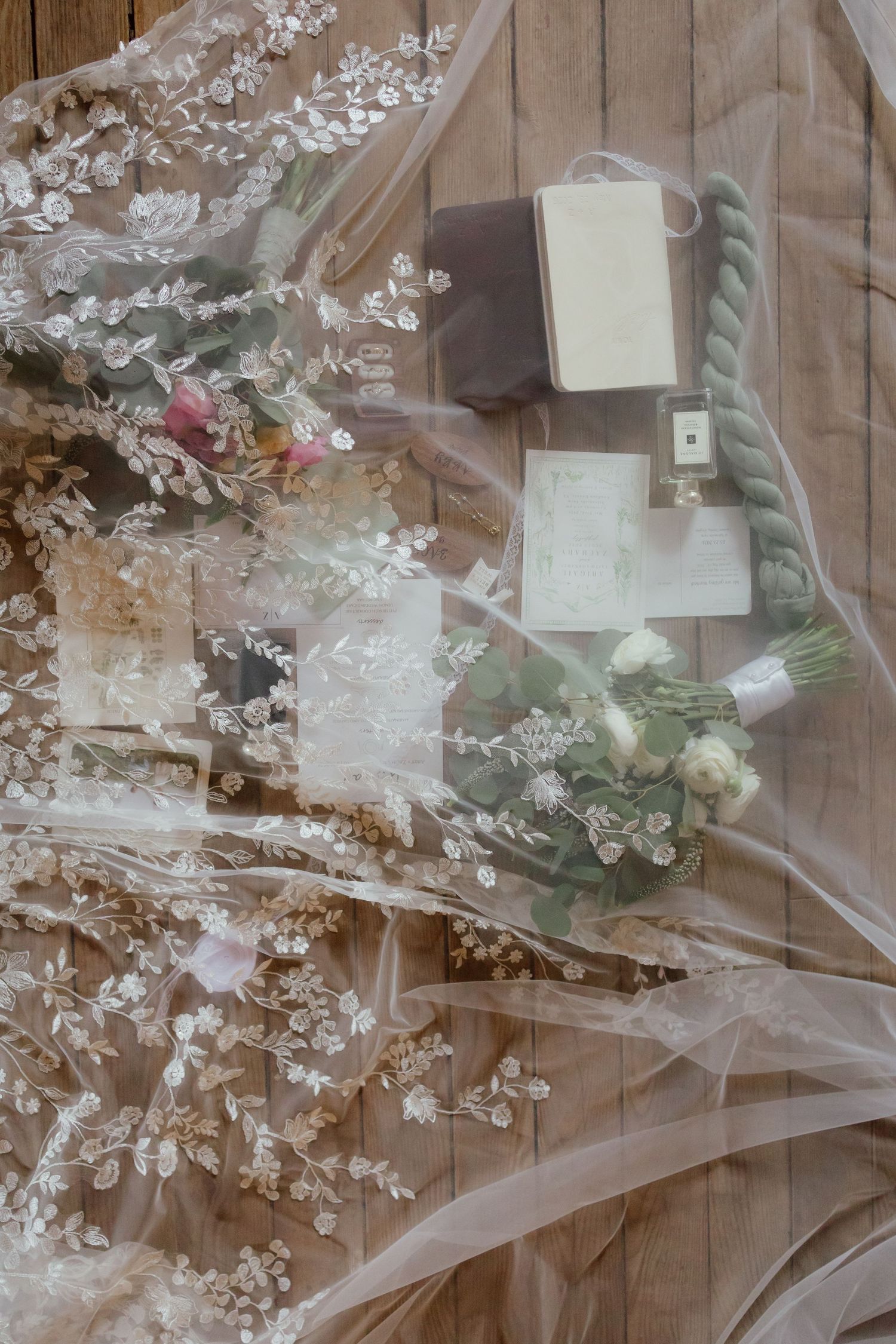 Flat lay of bridal details including lace veil, flowers, perfume, and stationery on wood floor.