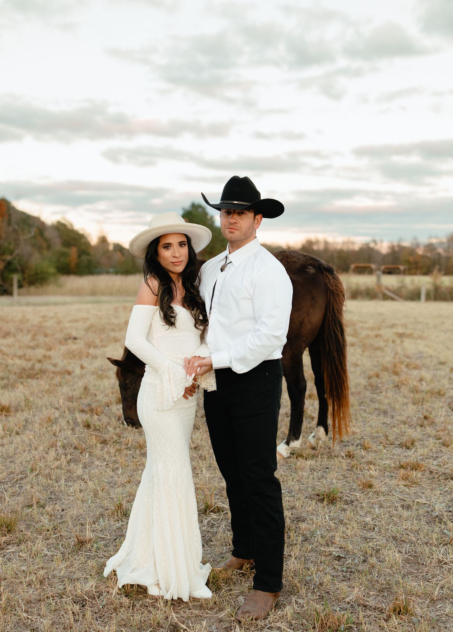 Bride in white lace gown and groom in black cowboy hat posing with horse on rustic farm.