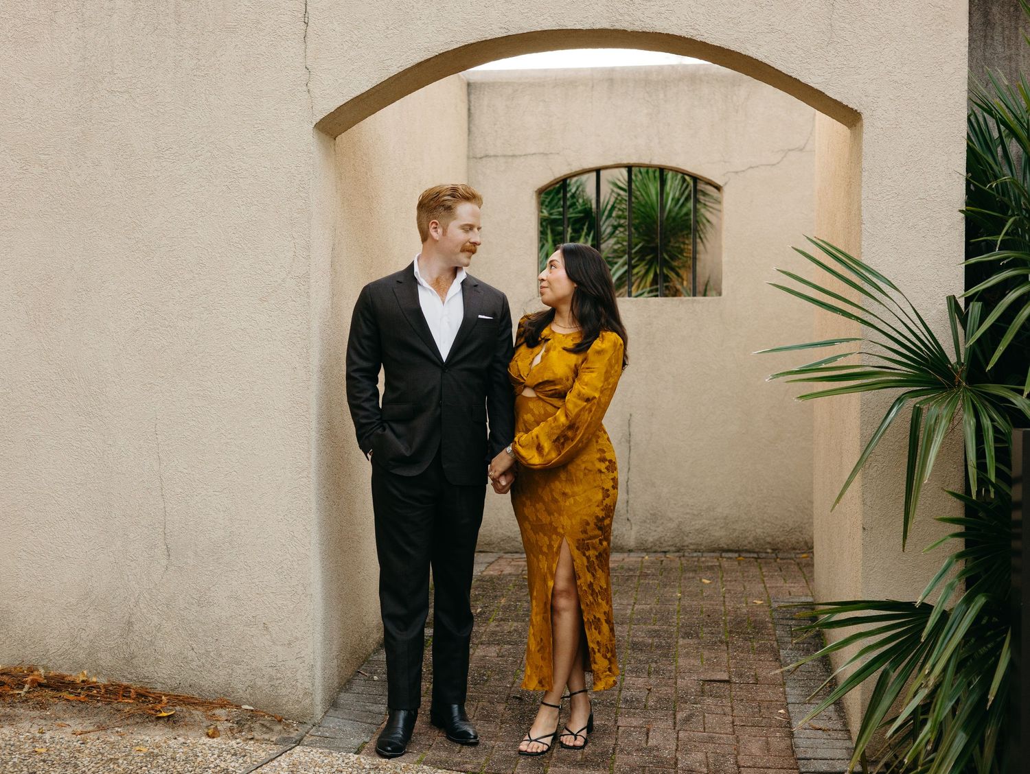Couple in elegant fall attire stand under stone arch, man in black suit, woman in gold dress, sharing a loving glance.