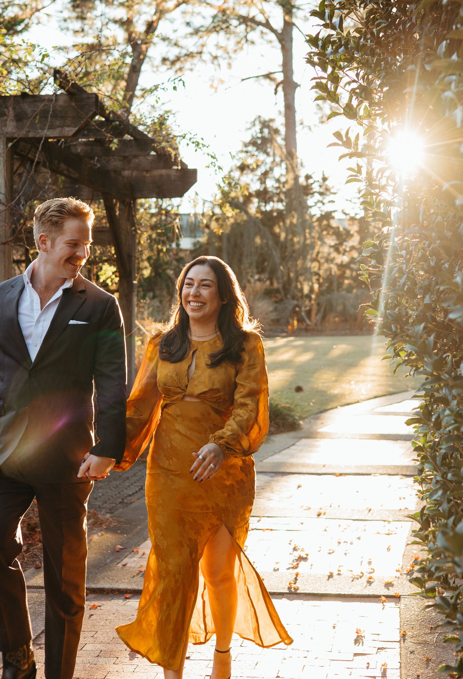 Happy couple walking hand in hand through sunlit garden path with golden lens flare.