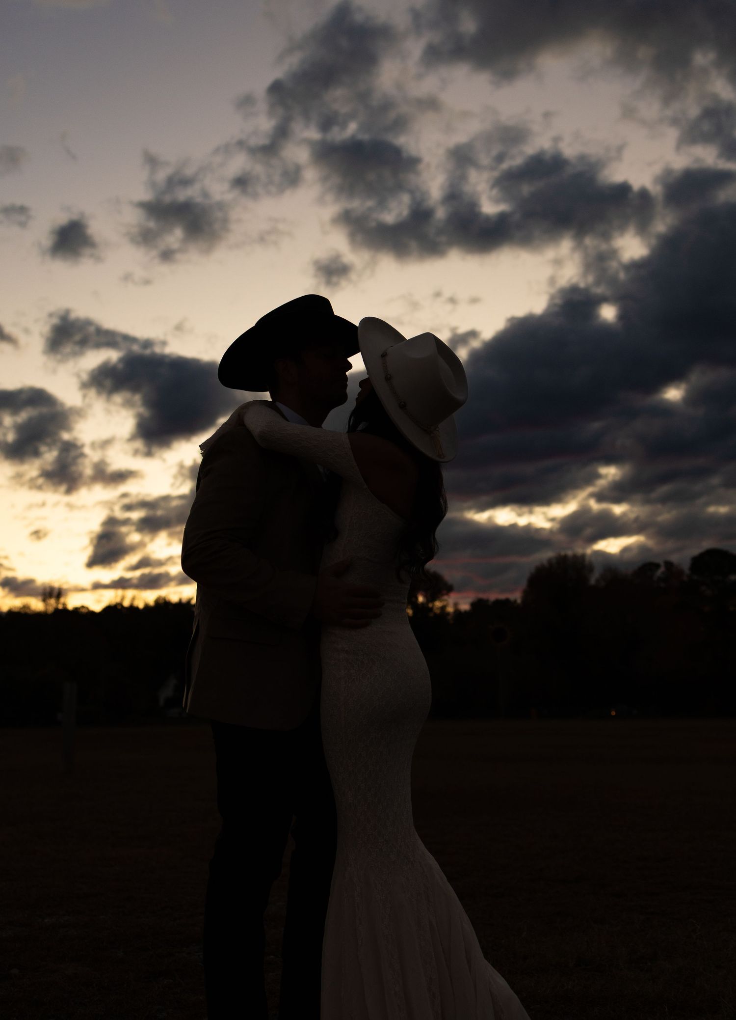 Romantic silhouette of bride and groom in cowboy hats embracing against dramatic sunset sky.