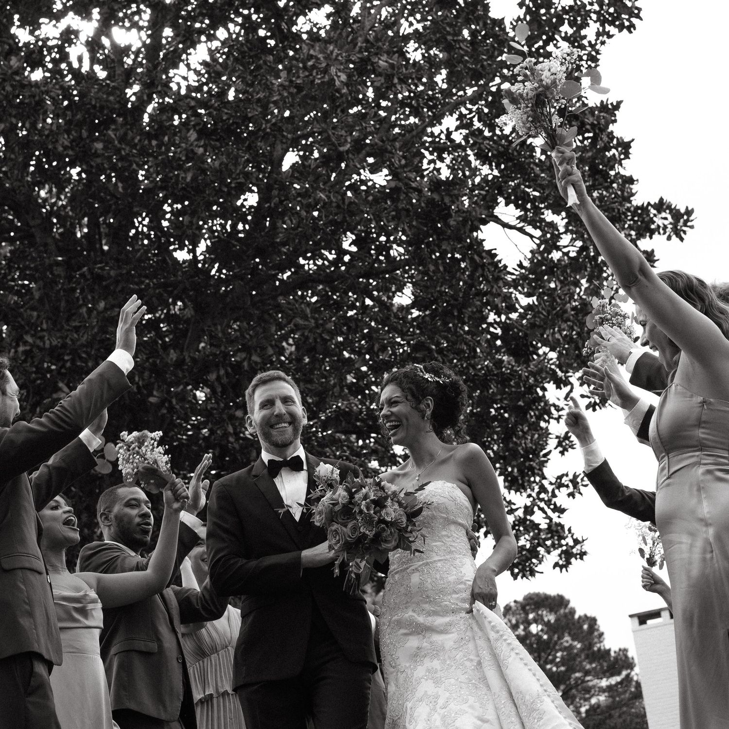 Black and white photo of smiling newlyweds walking through cheering wedding guests under a large tree.