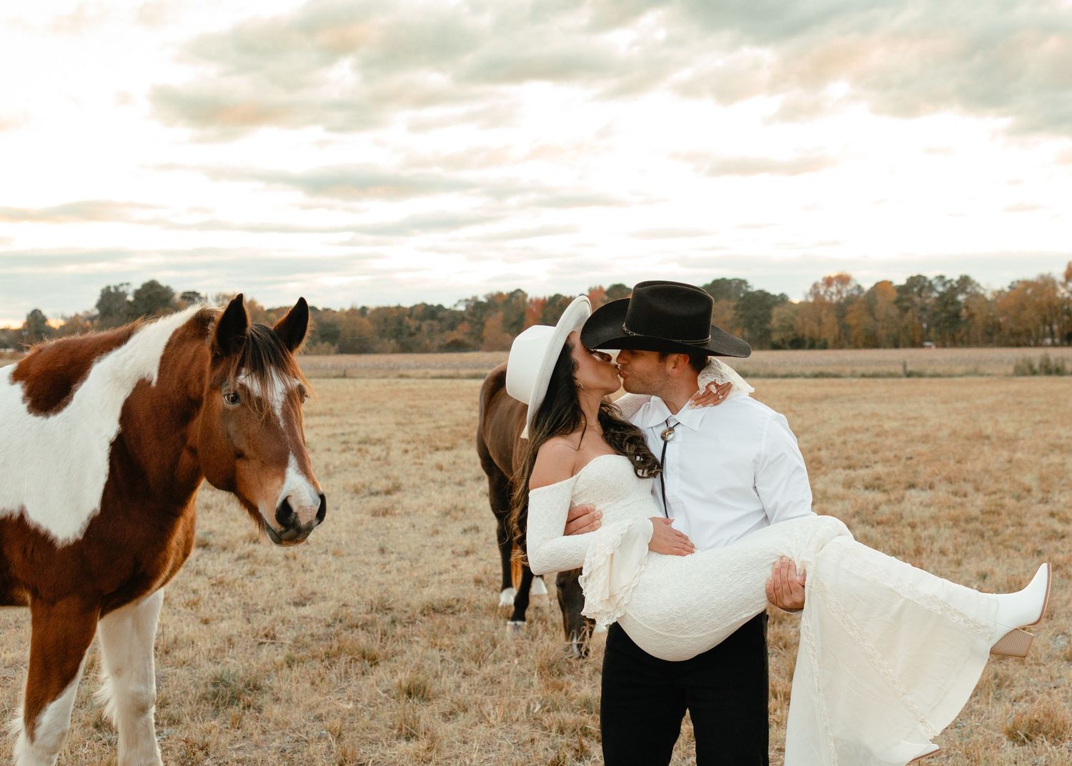 Bride and groom kissing near paint horses in an open field during golden hour at rustic country wedding.