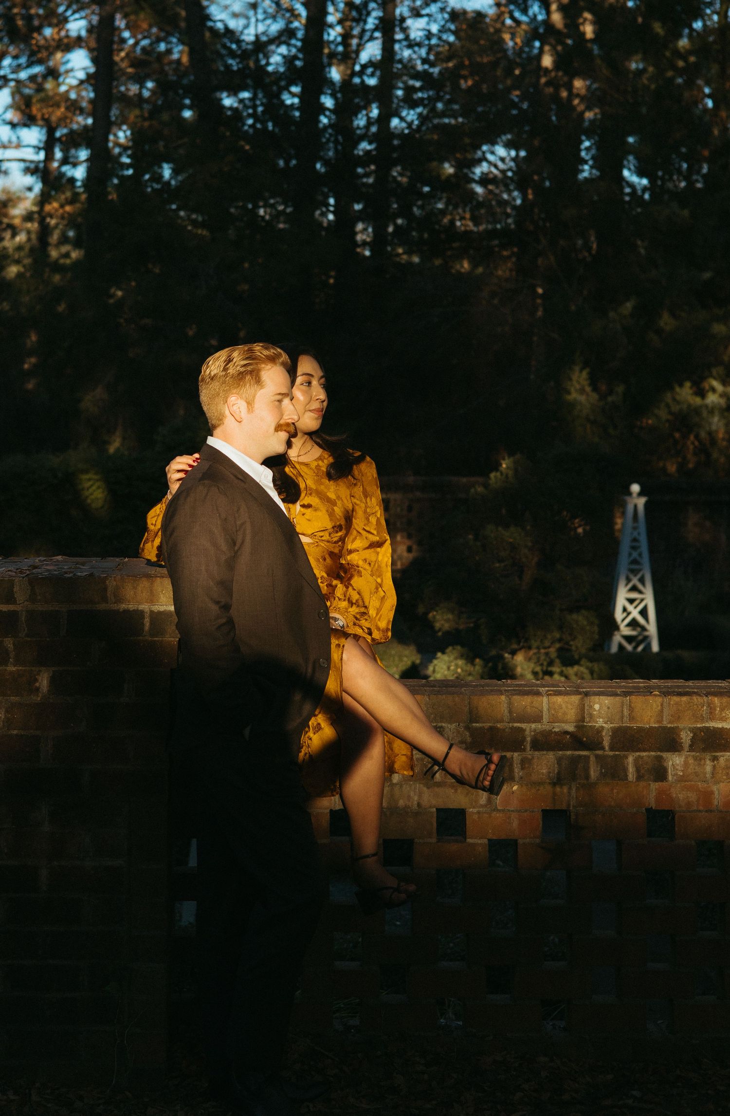Romantic couple posing at sunset by a brick wall in a garden, woman in gold dress.