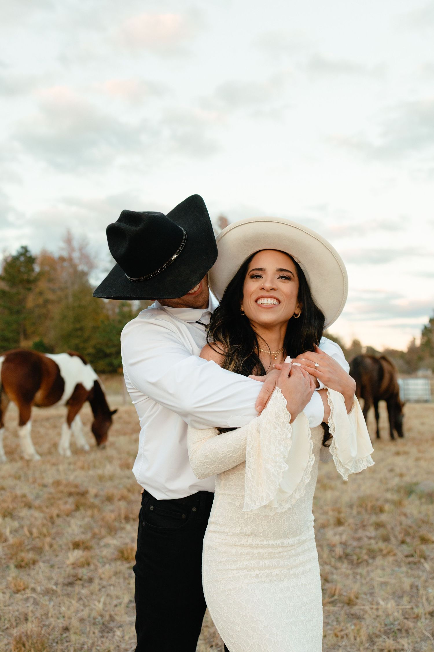 Couple in cowboy hats embracing in field with paint horses grazing in the background.