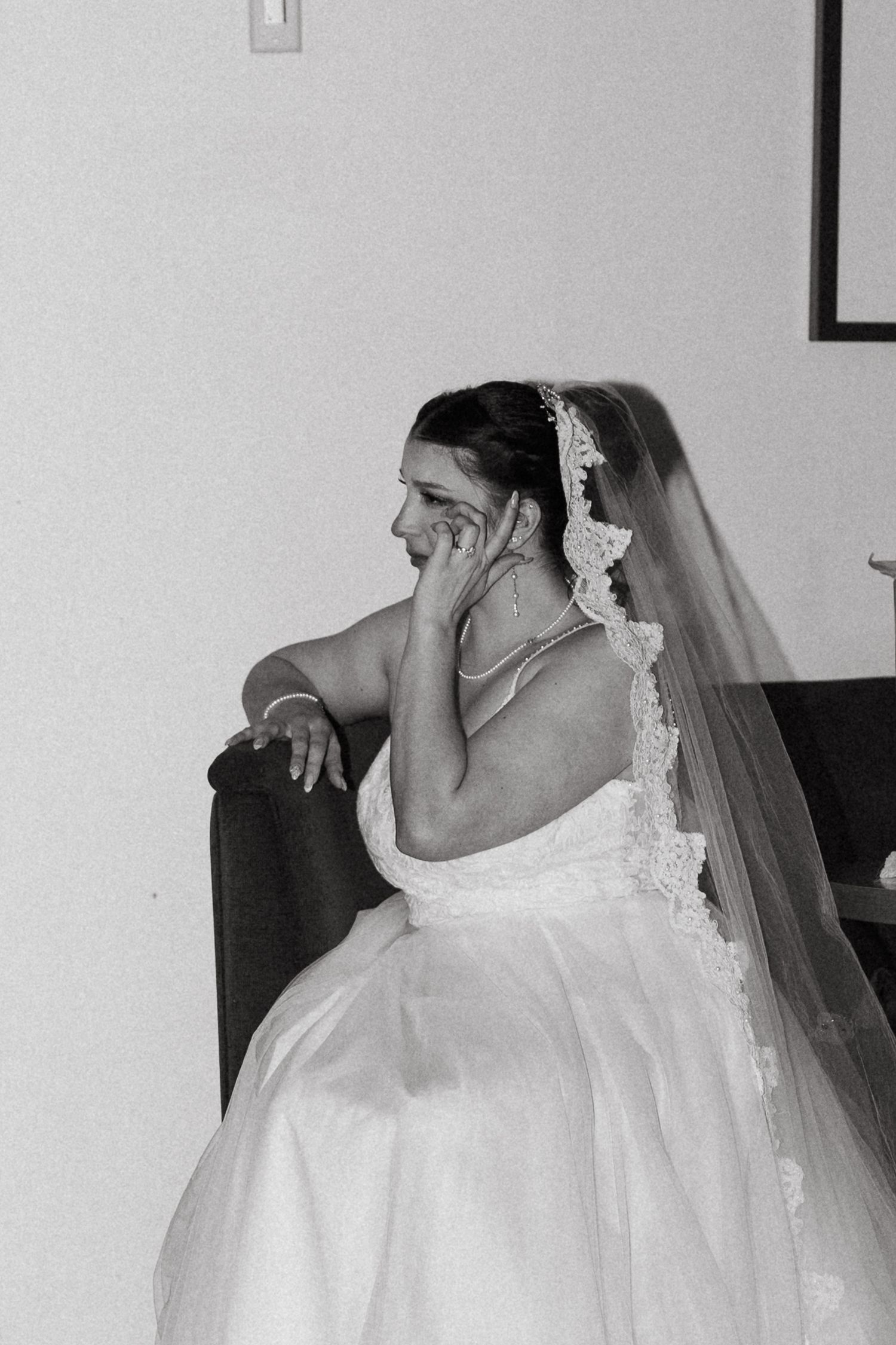 Emotional bride in white gown and lace veil sitting pensively in black and white wedding portrait.